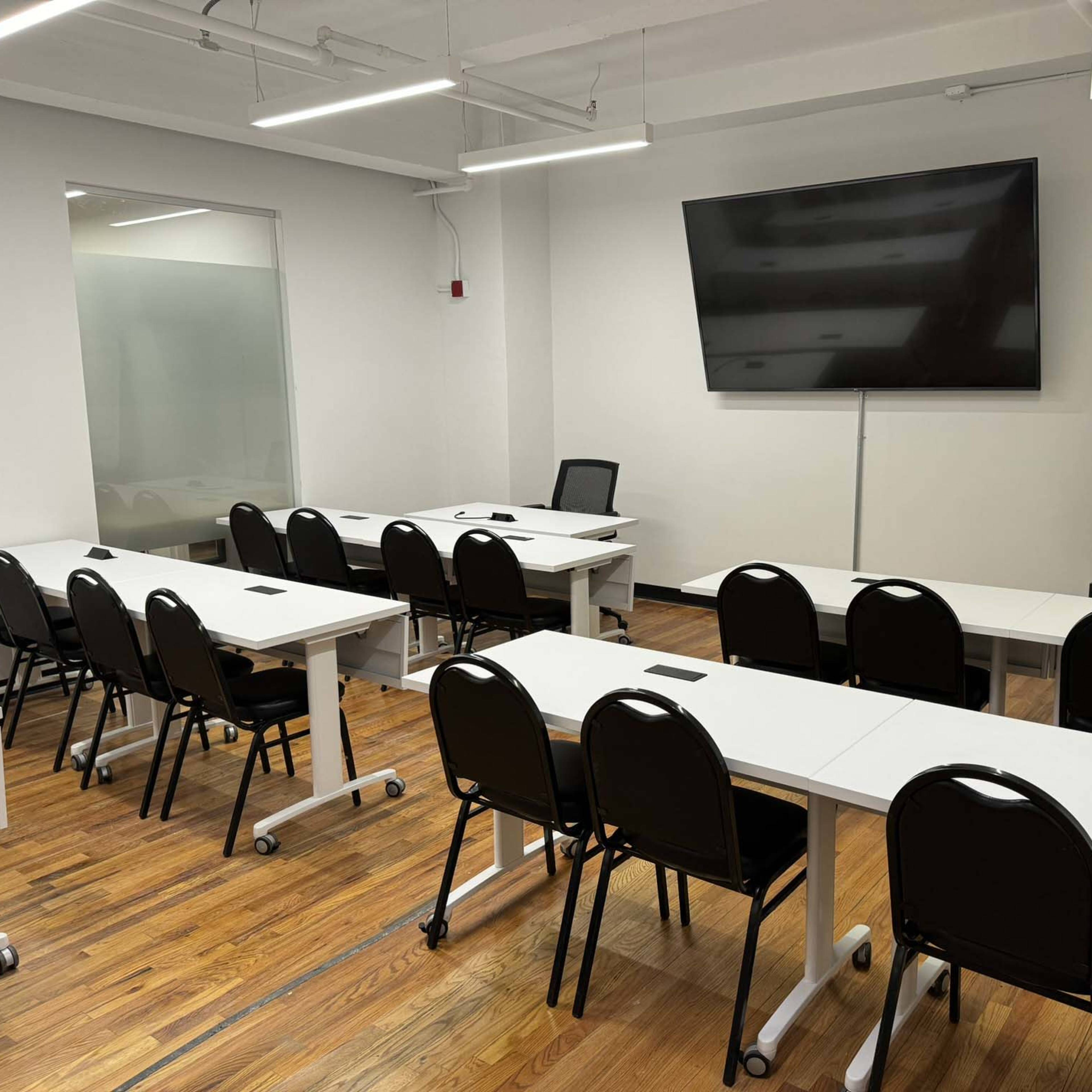 A modern classroom is set up with rows of tables and chairs, featuring a large wall-mounted screen and a whiteboard at the front.