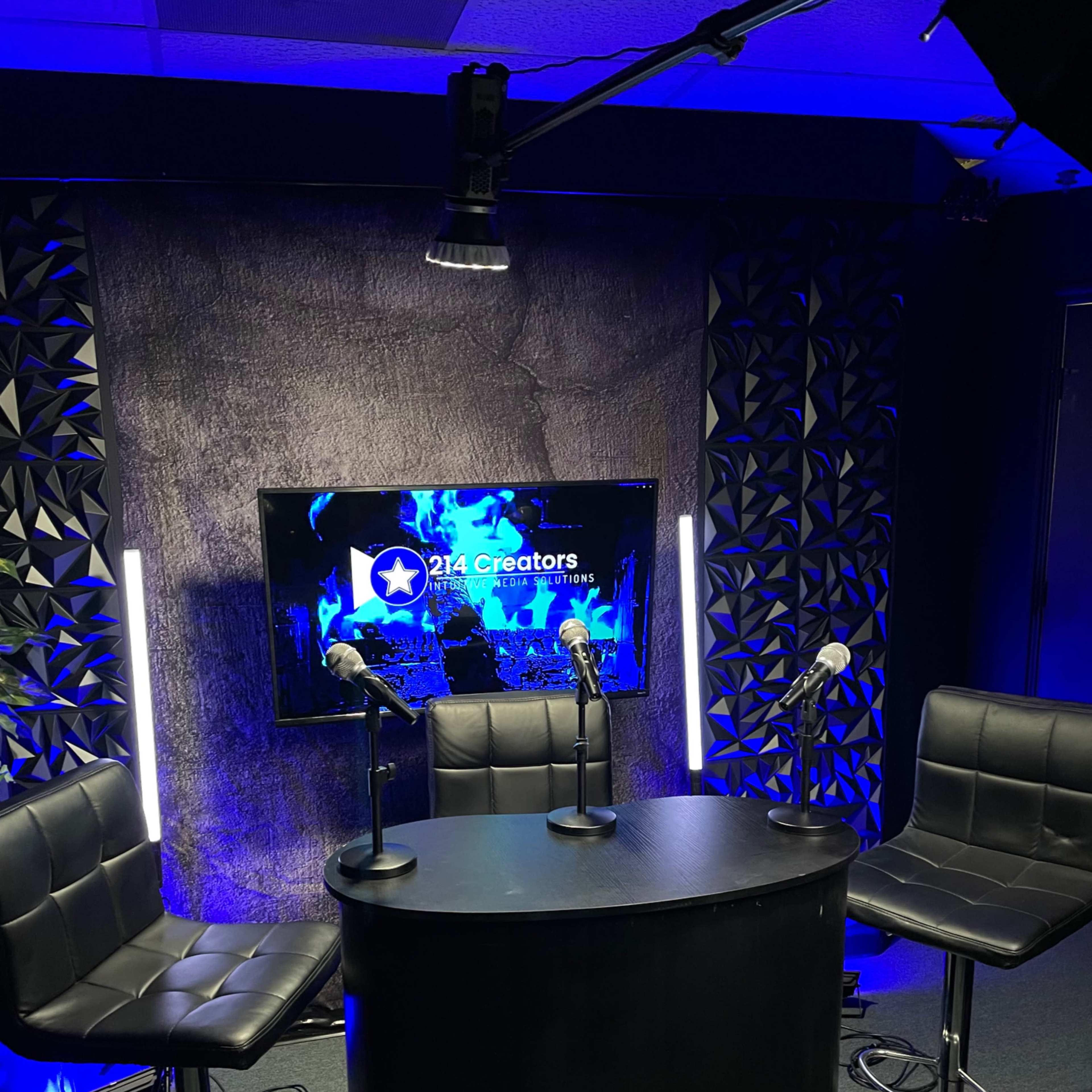 A modern studio setup with three barstools and a circular table in front of a backdrop featuring a screen that displays "214 Creators."