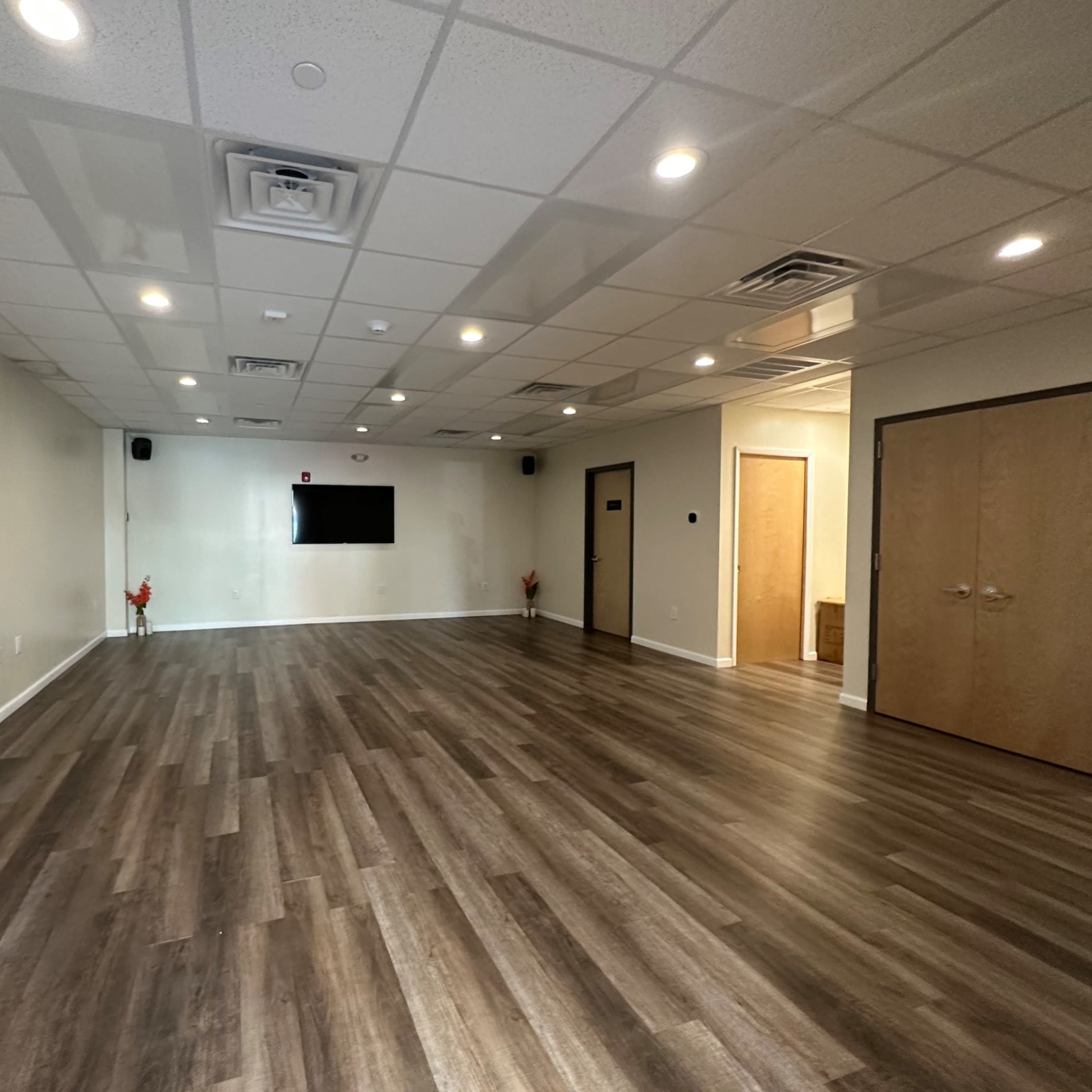 A spacious, empty room with hardwood flooring, a flat-screen TV mounted on the wall, and two closed doors.