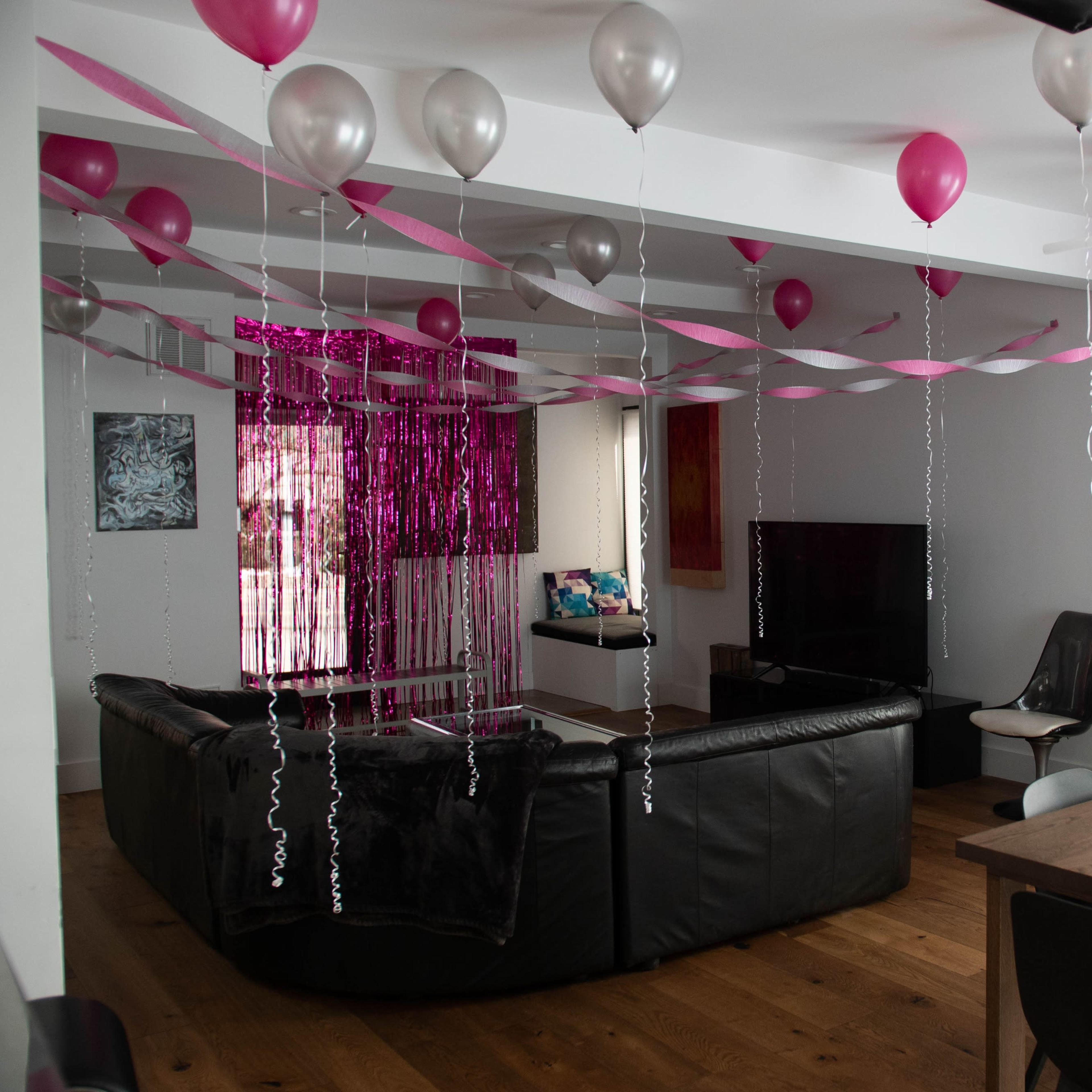 The living room is decorated with pink and silver balloons and streamers hanging from the ceiling, creating a festive atmosphere.