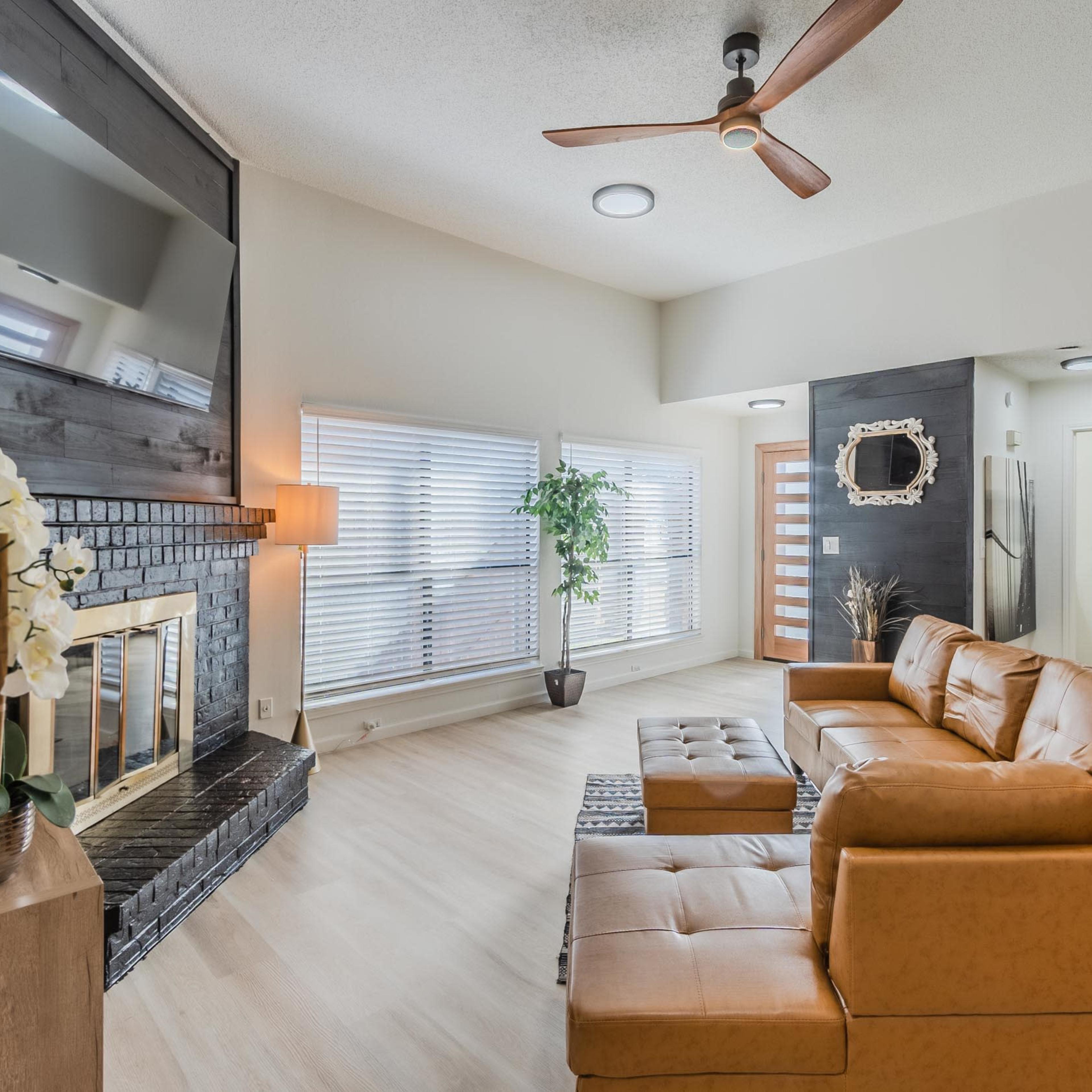A modern living room features a tan leather sectional sofa, a fireplace with a black surround, and large windows with blinds.