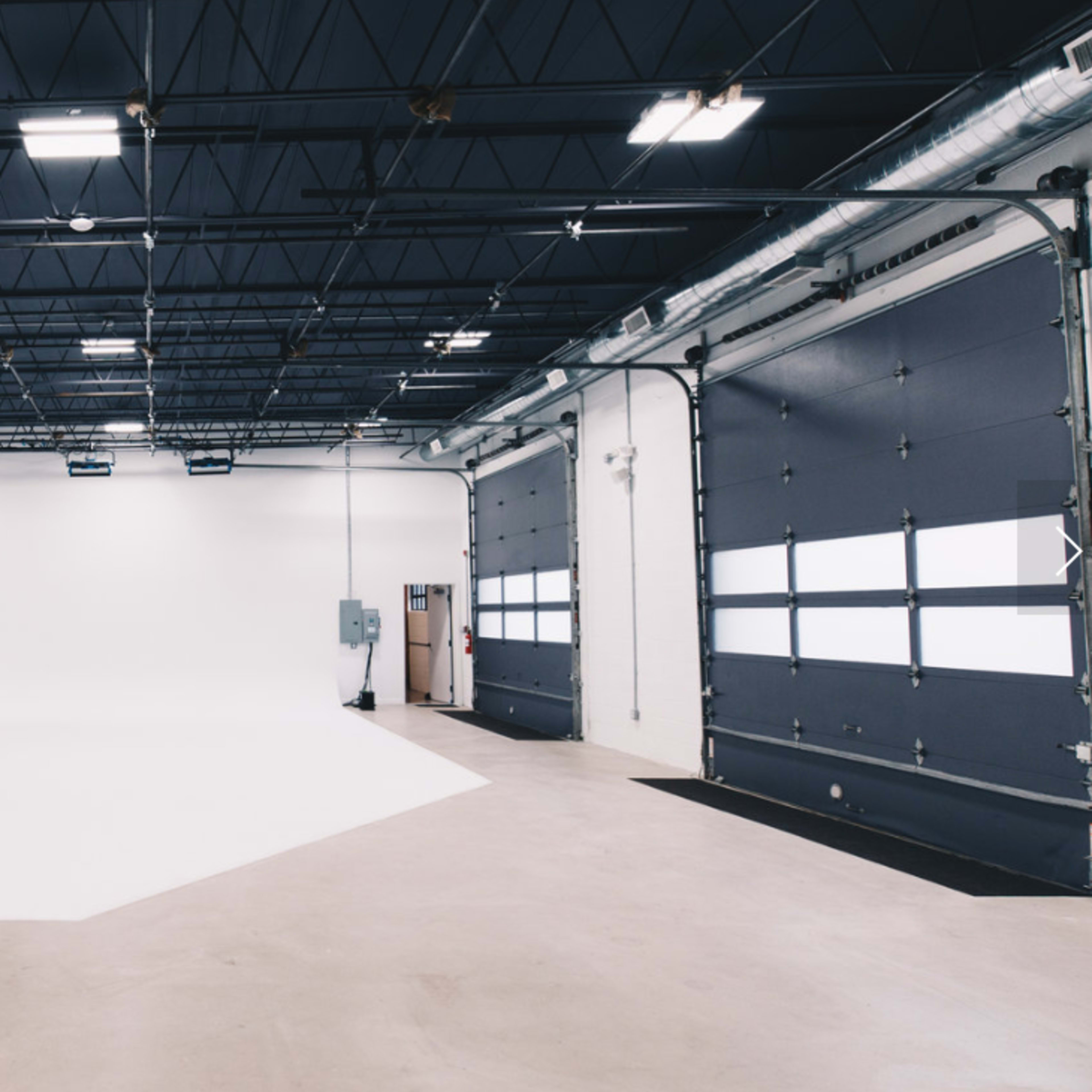 A large, empty studio space features a white backdrop and industrial lighting, with a pair of overhead doors on one side.