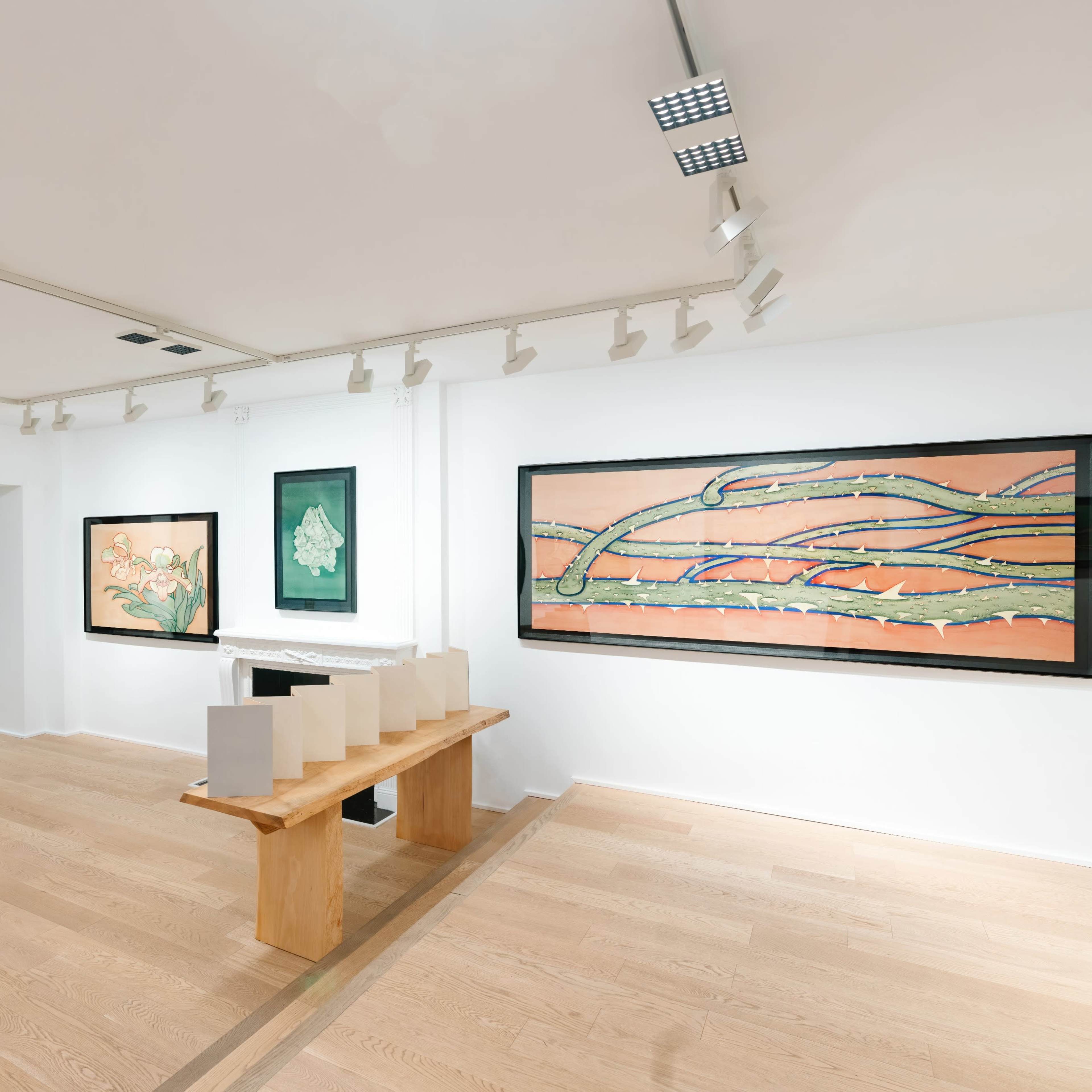 A modern art gallery features several large framed paintings on white walls, with a wooden table displaying white objects in the center of the room.