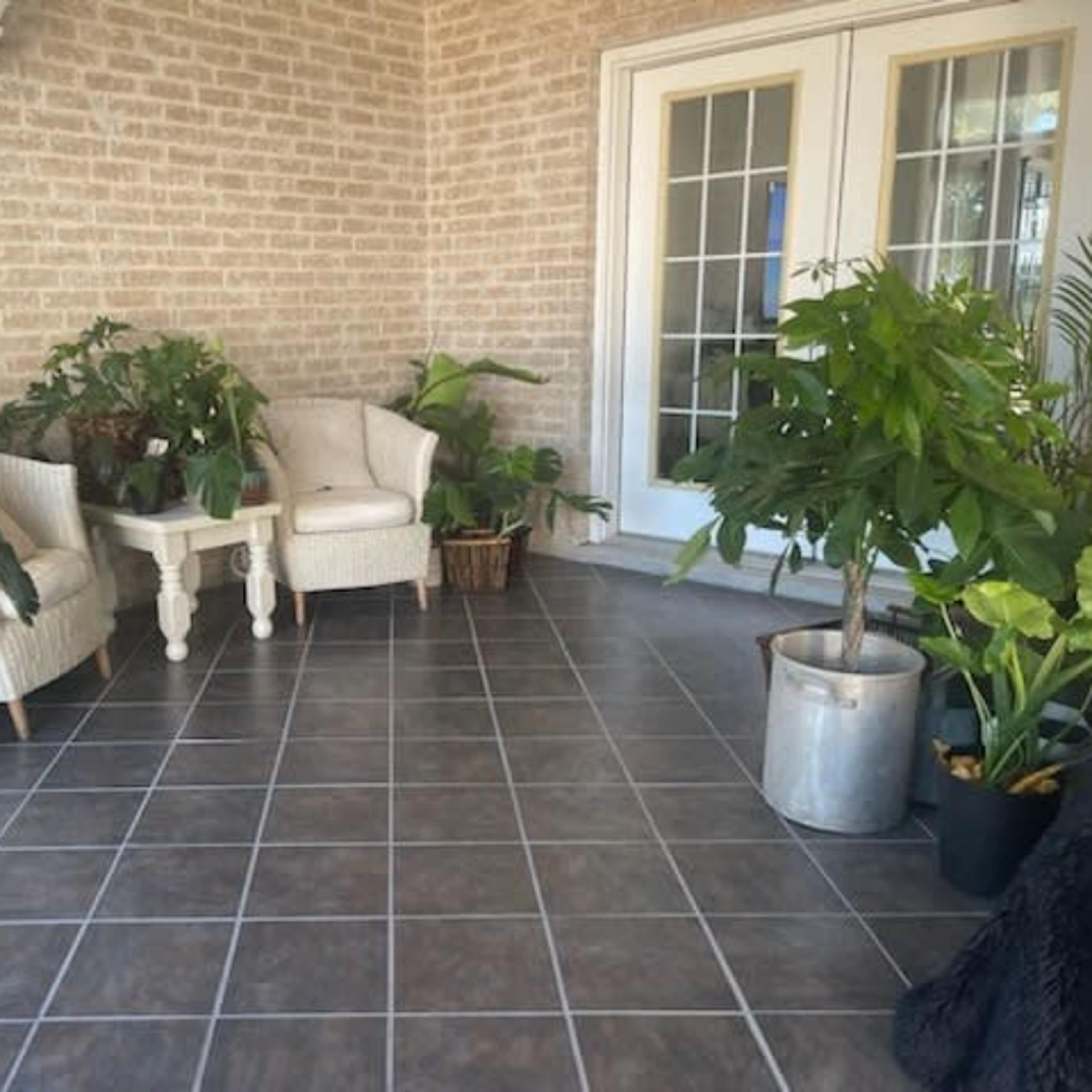 A patio area features two white armchairs, a small table, and various potted plants on a tiled floor.