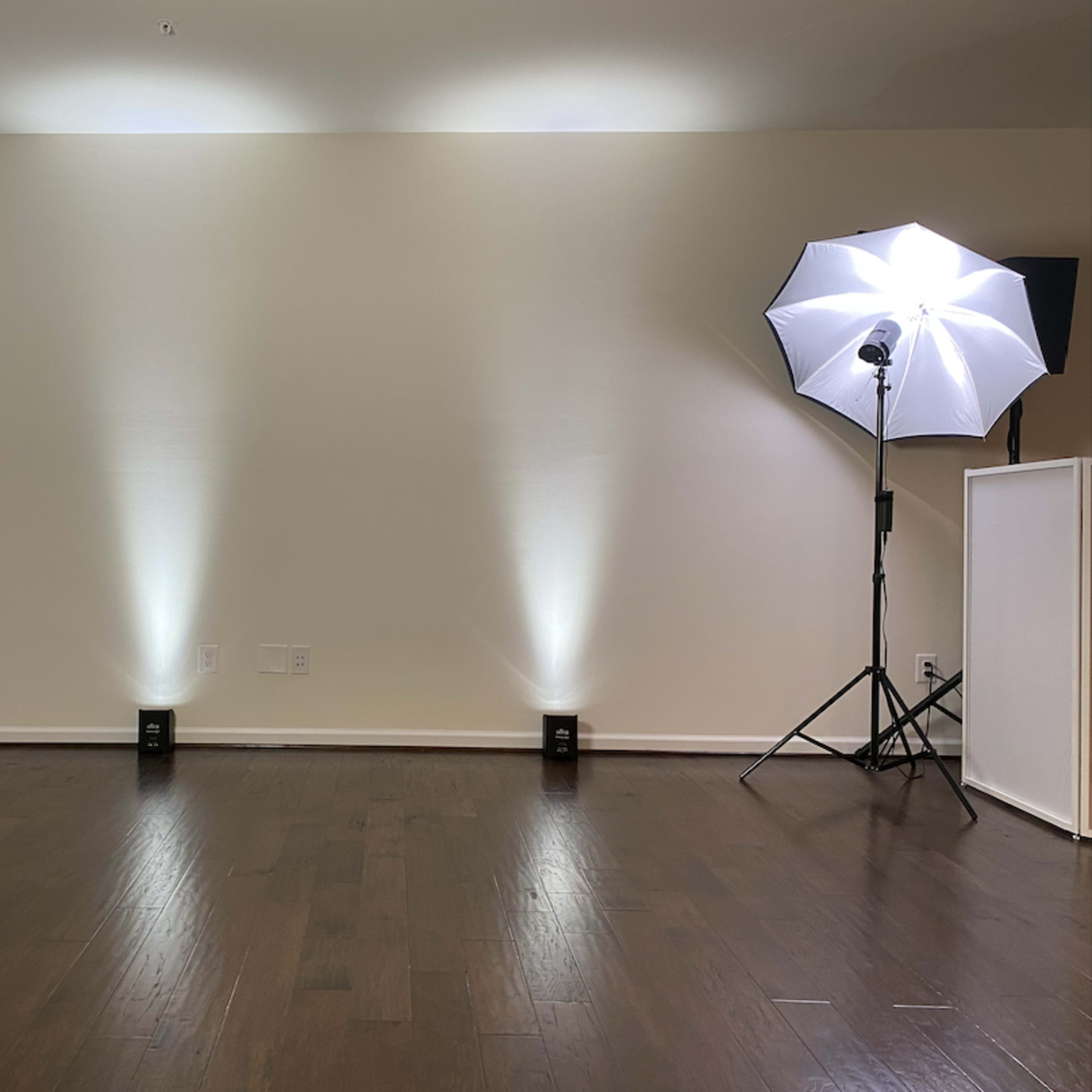 A photography studio setting with two light sources, one softbox and one strobe light, positioned in a room with hardwood flooring and a plain wall.