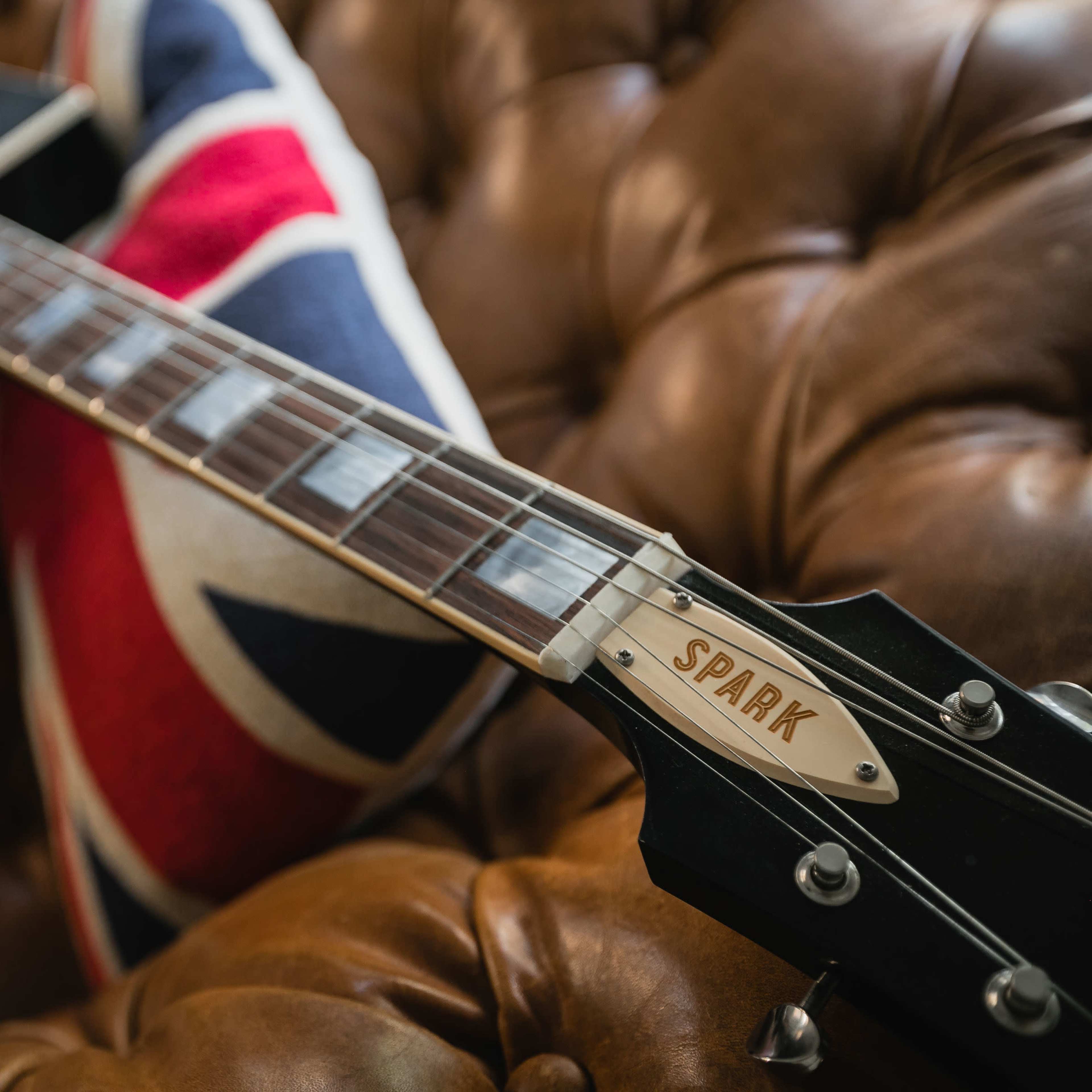 A black electric guitar with the name "SPARK" on the headstock rests on a brown leather couch adorned with a Union Jack pillow.