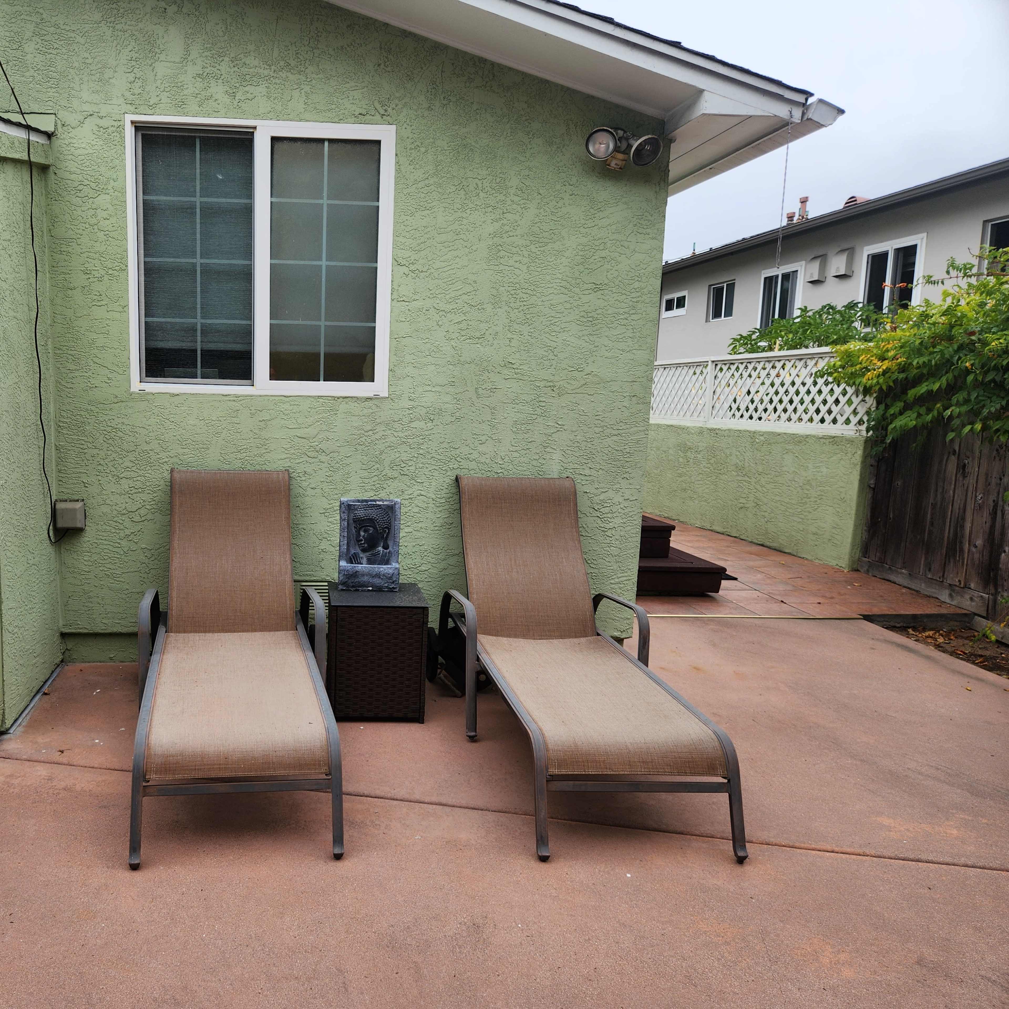 Two lounge chairs positioned next to a gray stone fountain against a green exterior wall of a house.