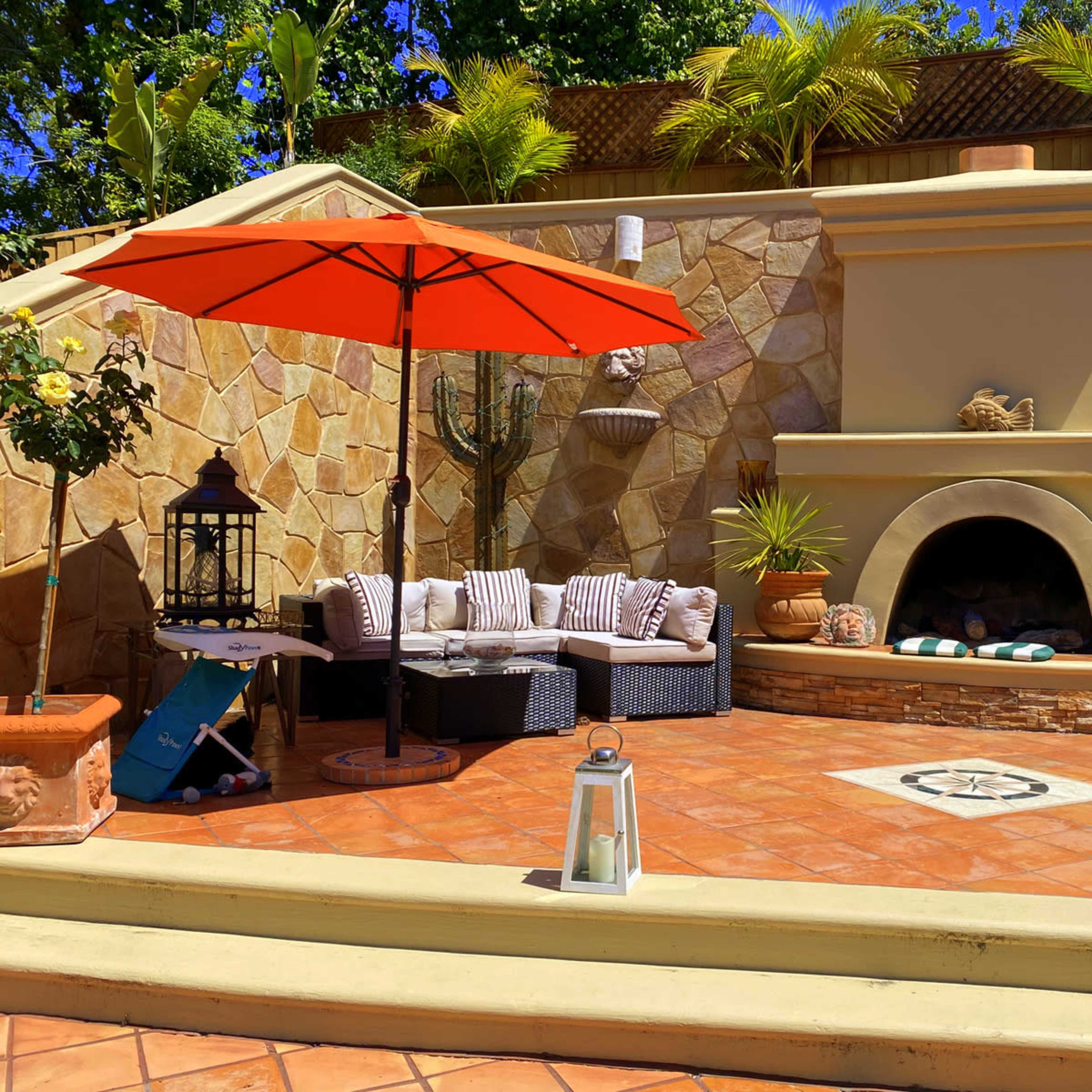 A landscaped outdoor seating area with a stone fireplace, a circular decorative tile design, a large orange umbrella, and various potted plants.