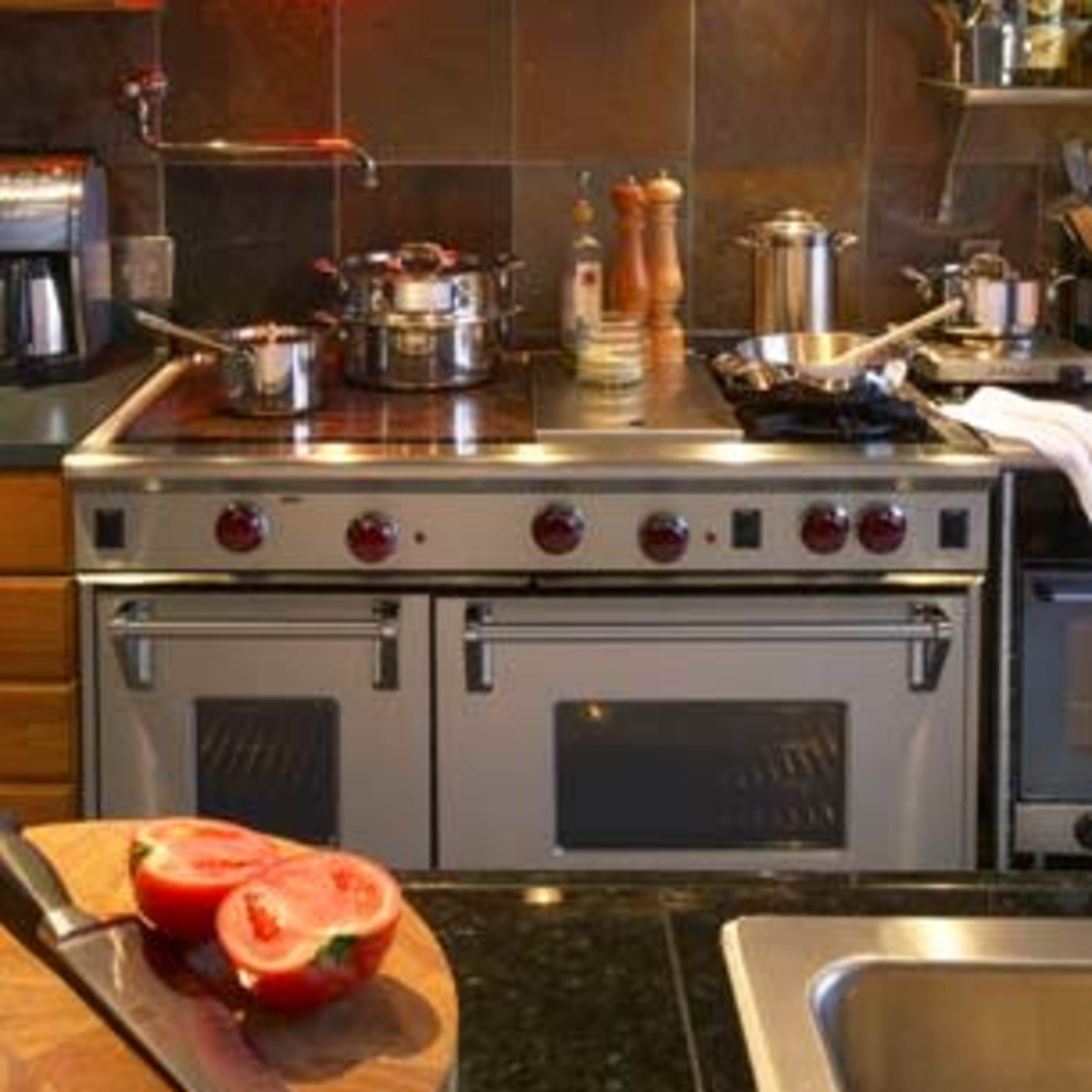A modern kitchen with a stainless steel stove, several pots on the burners, and a chopping board with sliced tomatoes in the foreground.