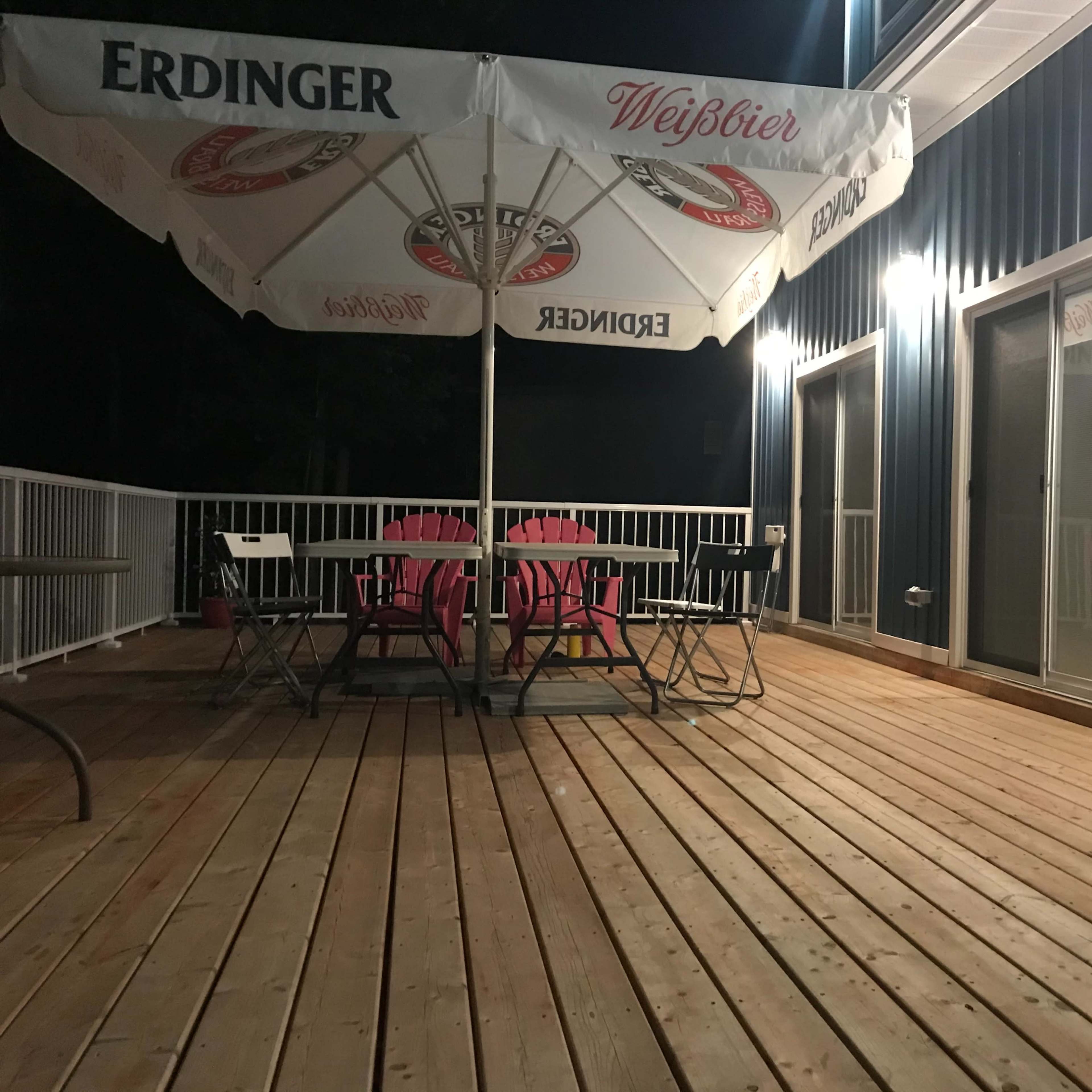 A wooden deck at night with a large umbrella and a table surrounded by chairs, illuminated by nearby lights.