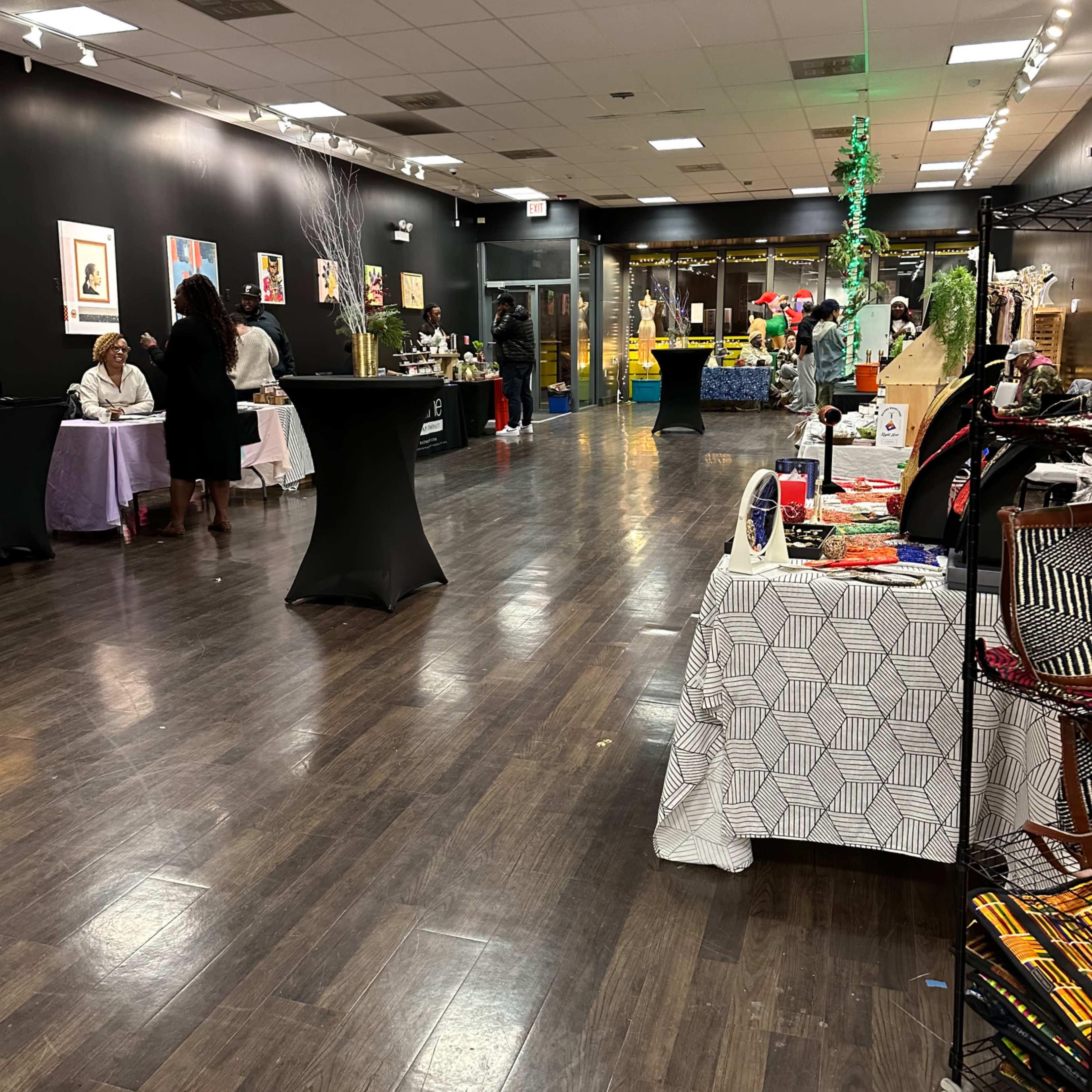 An indoor venue with people gathered around tables displaying various items for sale, set against a backdrop of artwork on the walls.