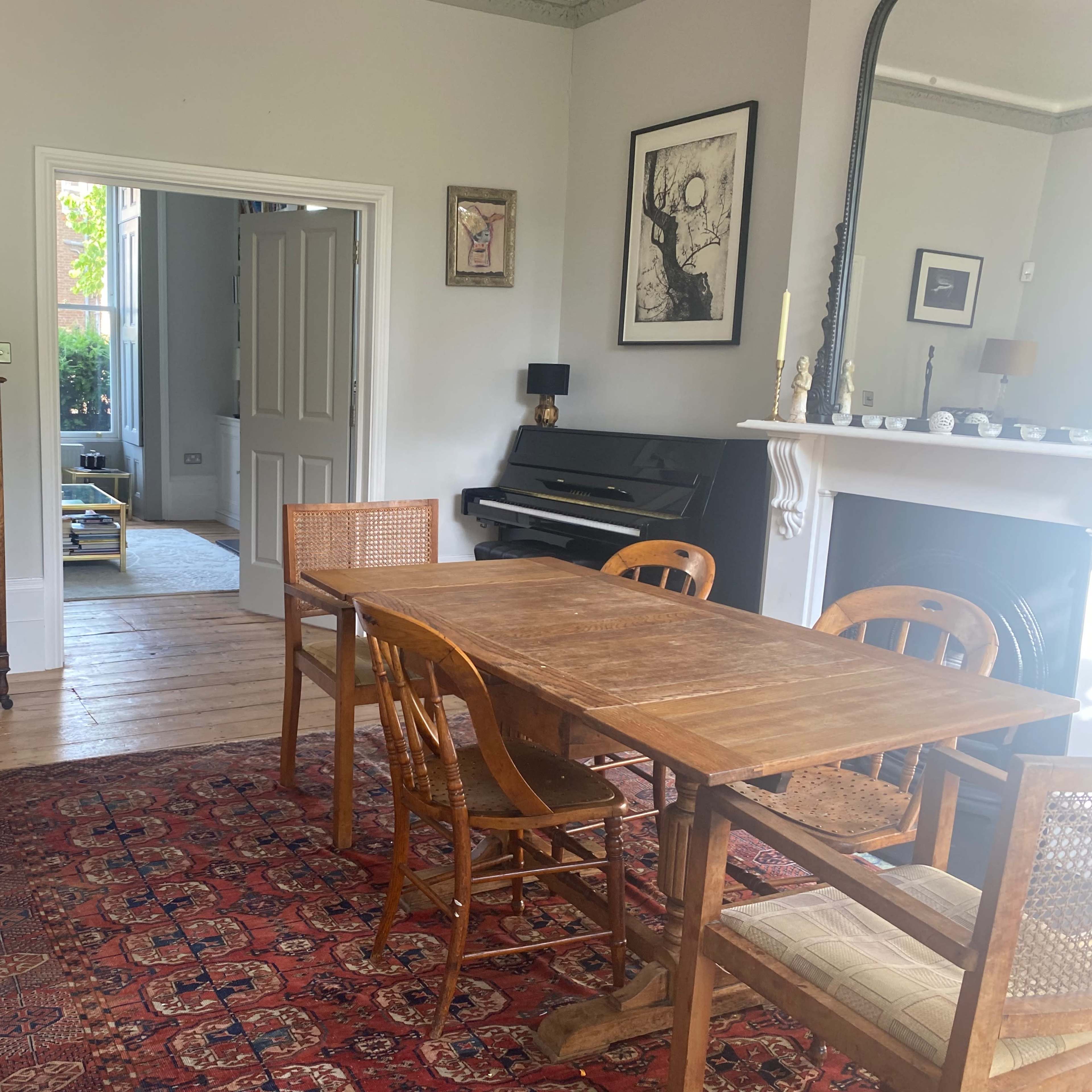 A room with a wooden dining table and chairs, an antique dresser, a piano, and a fireplace, complemented by artwork on the walls.