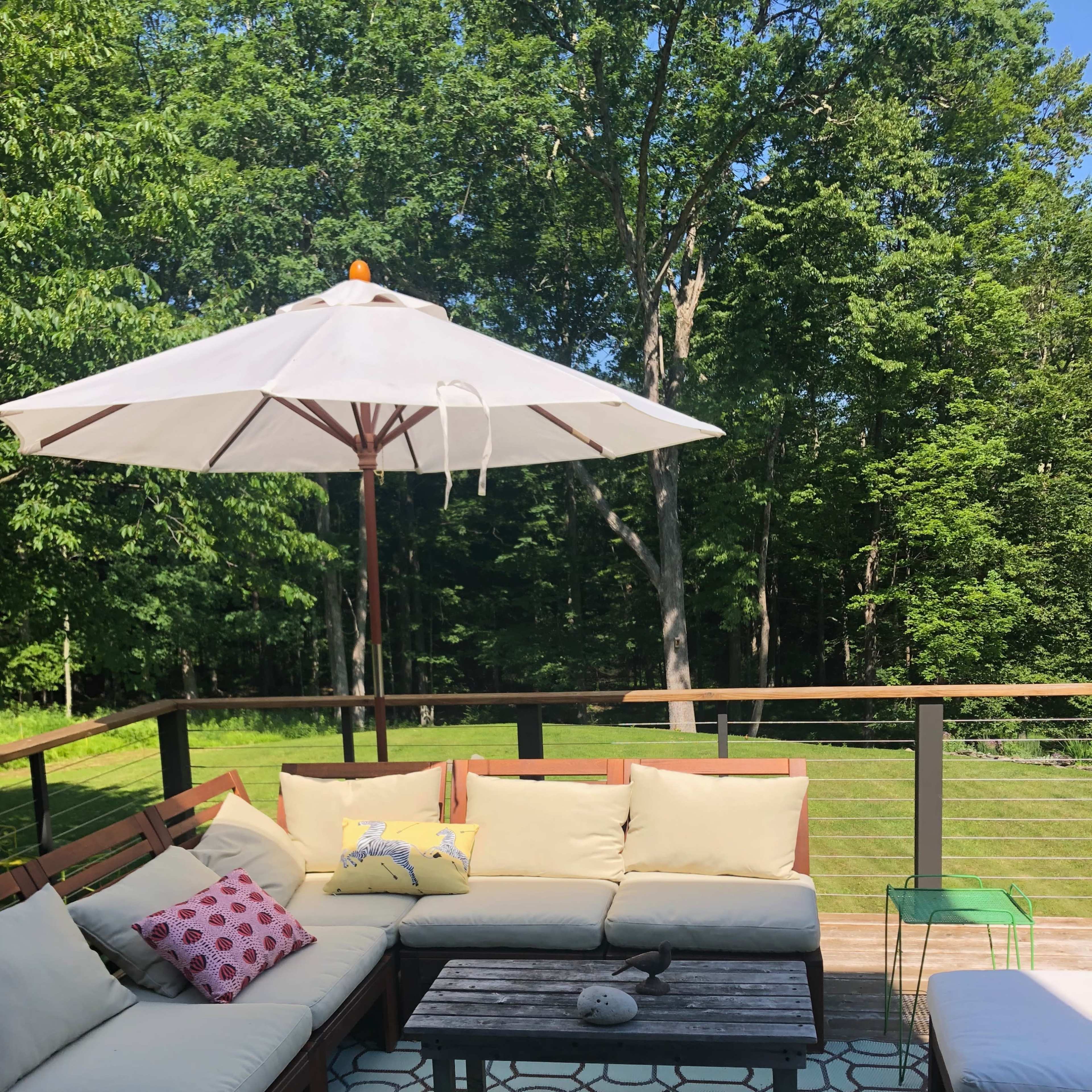 A furnished outdoor seating area features a sectional couch with cushions, a coffee table, and a large umbrella on a deck surrounded by greenery.