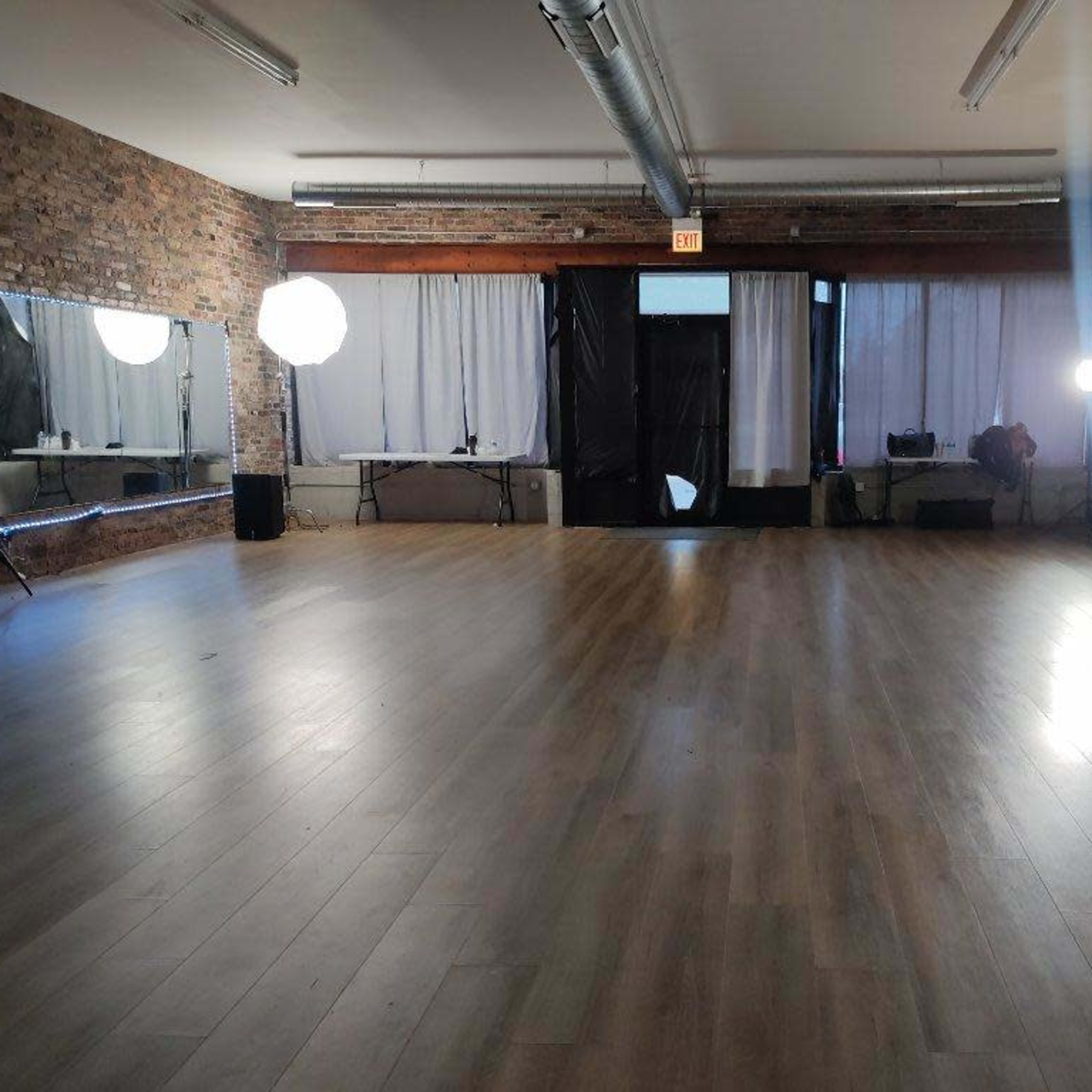 A spacious empty studio with wooden flooring, exposed brick walls, and large windows covered by white curtains, illuminated by two softbox lights.