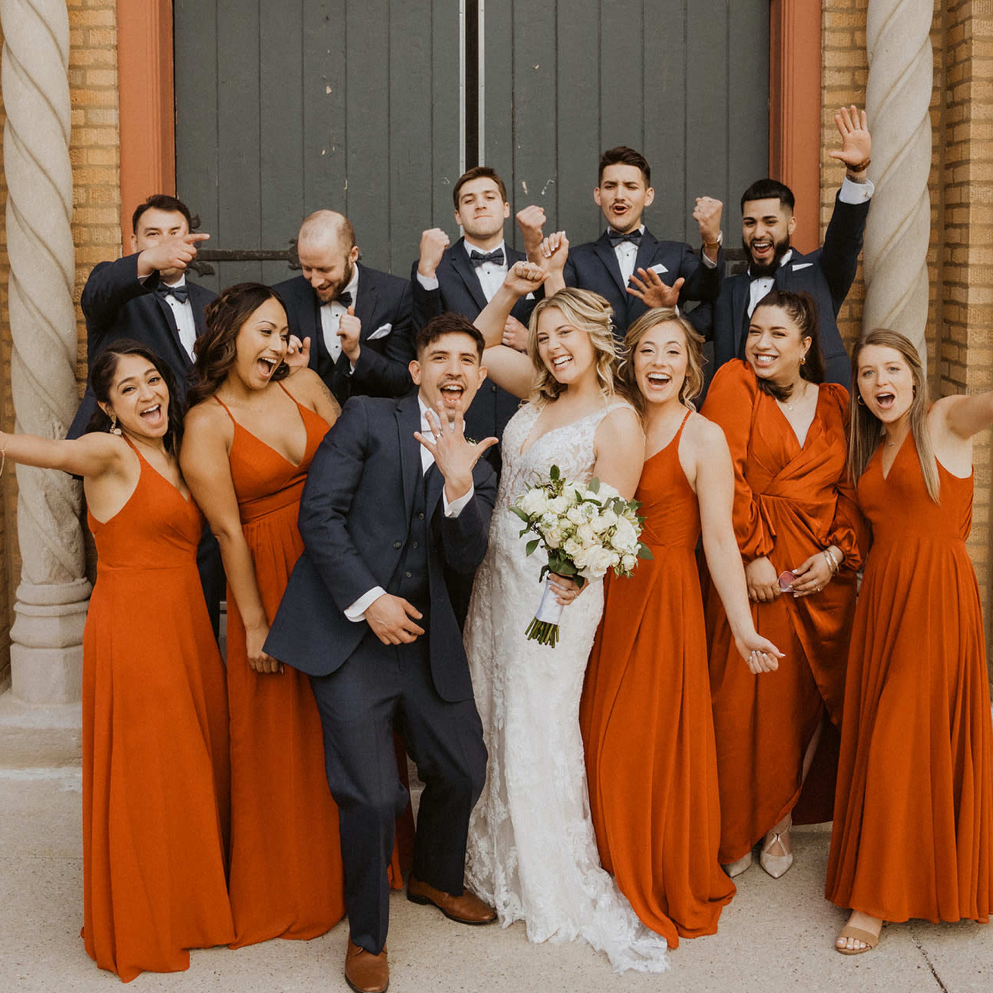 A wedding party poses joyfully in front of a large door, wearing coordinated attire with the bride in a white gown and bouquet.