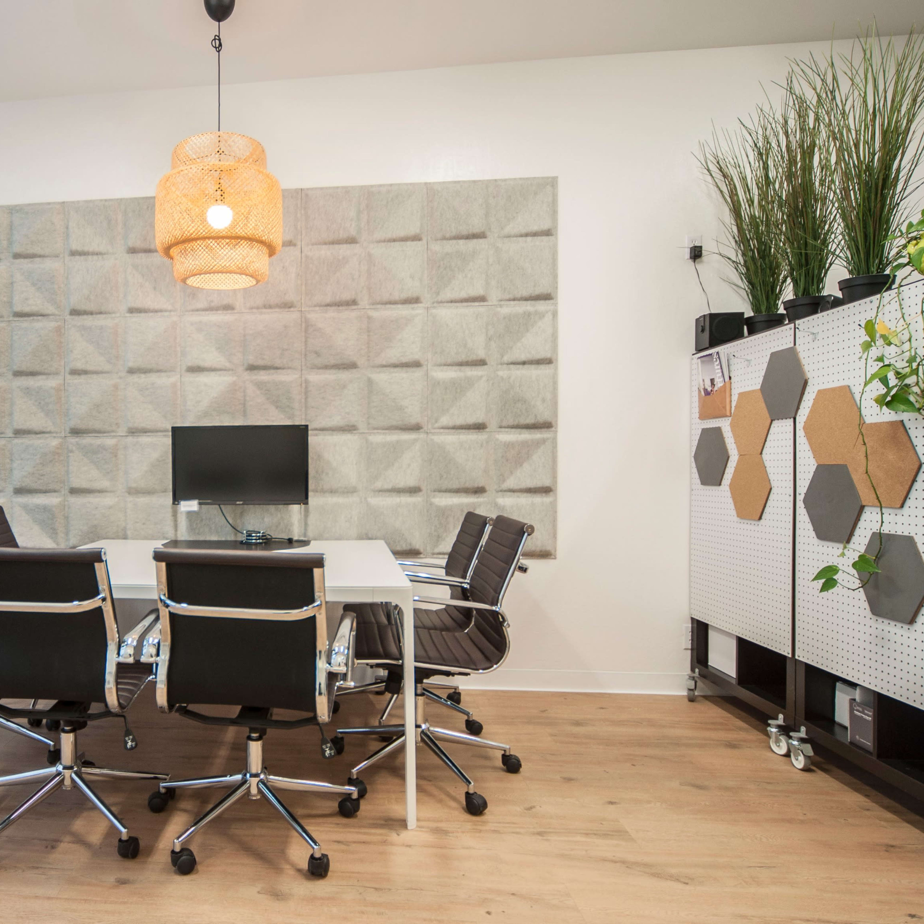 A modern conference room features a round white table surrounded by black swivel chairs, with a wall-mounted monitor and a large plant near a textured gray wall.