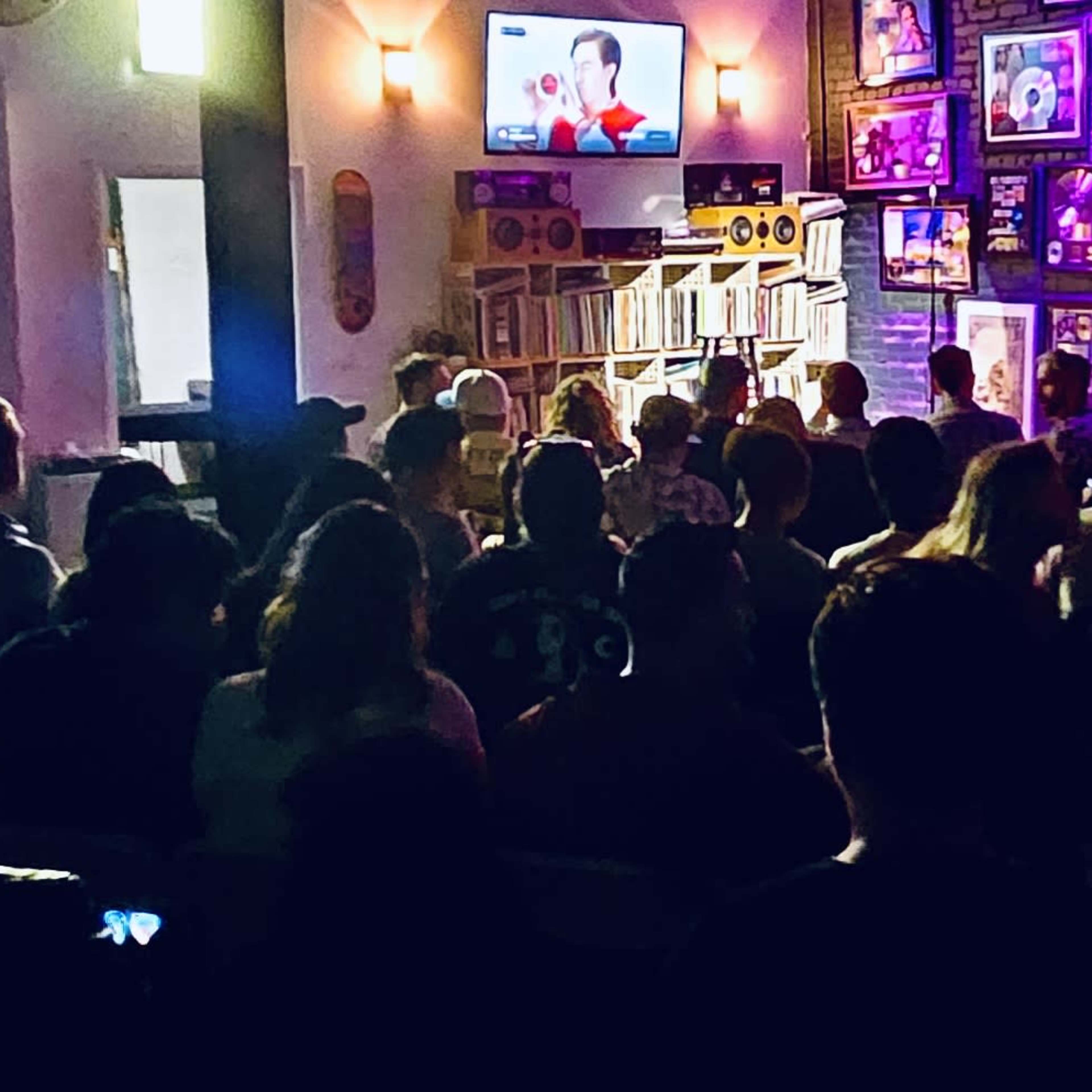 A large audience sits in a dimly lit room, facing a stage adorned with music awards and a screen displaying a visual presentation.
