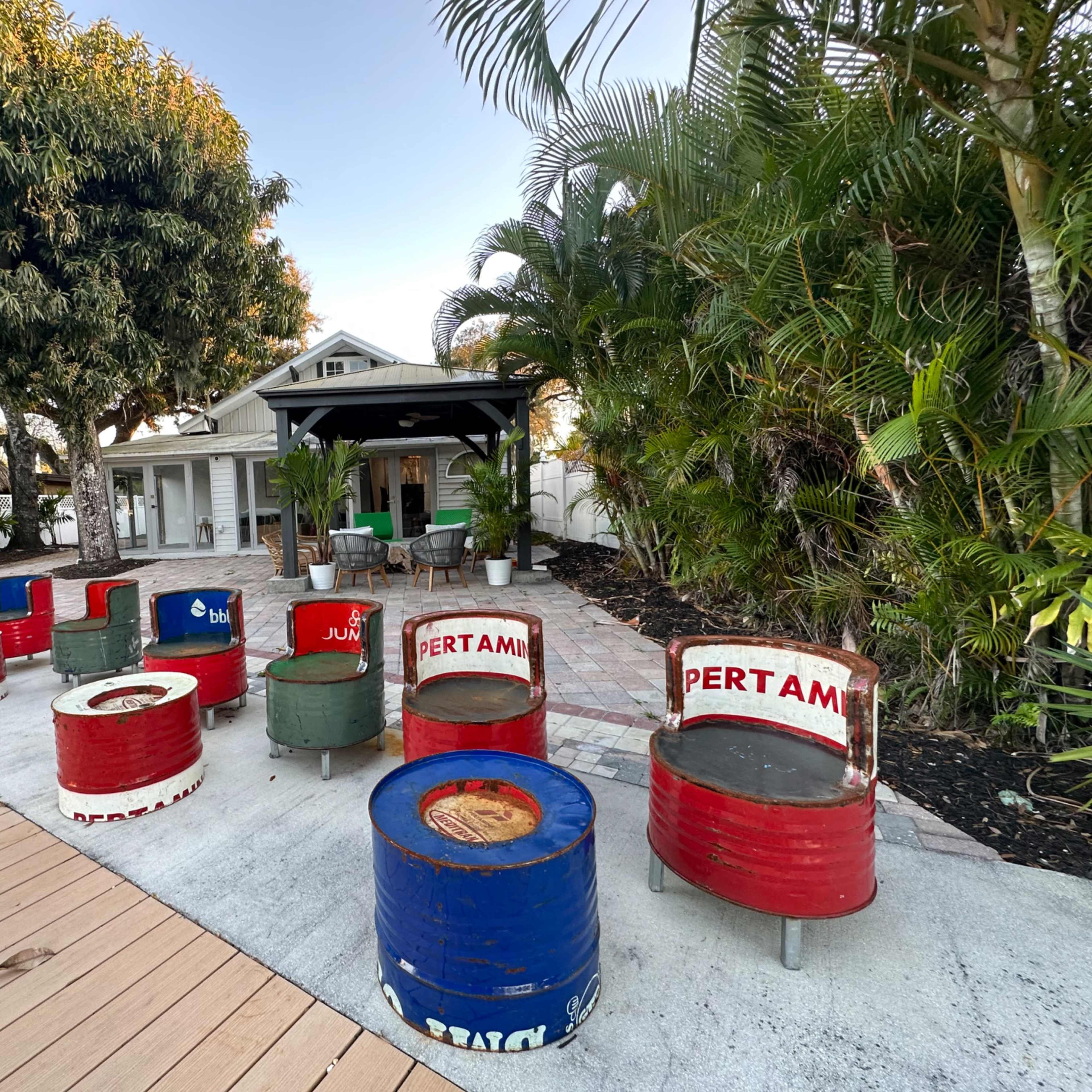 The scene shows a seating area made from colorful oil drums arranged in a circular pattern near a tropical house and palm trees.