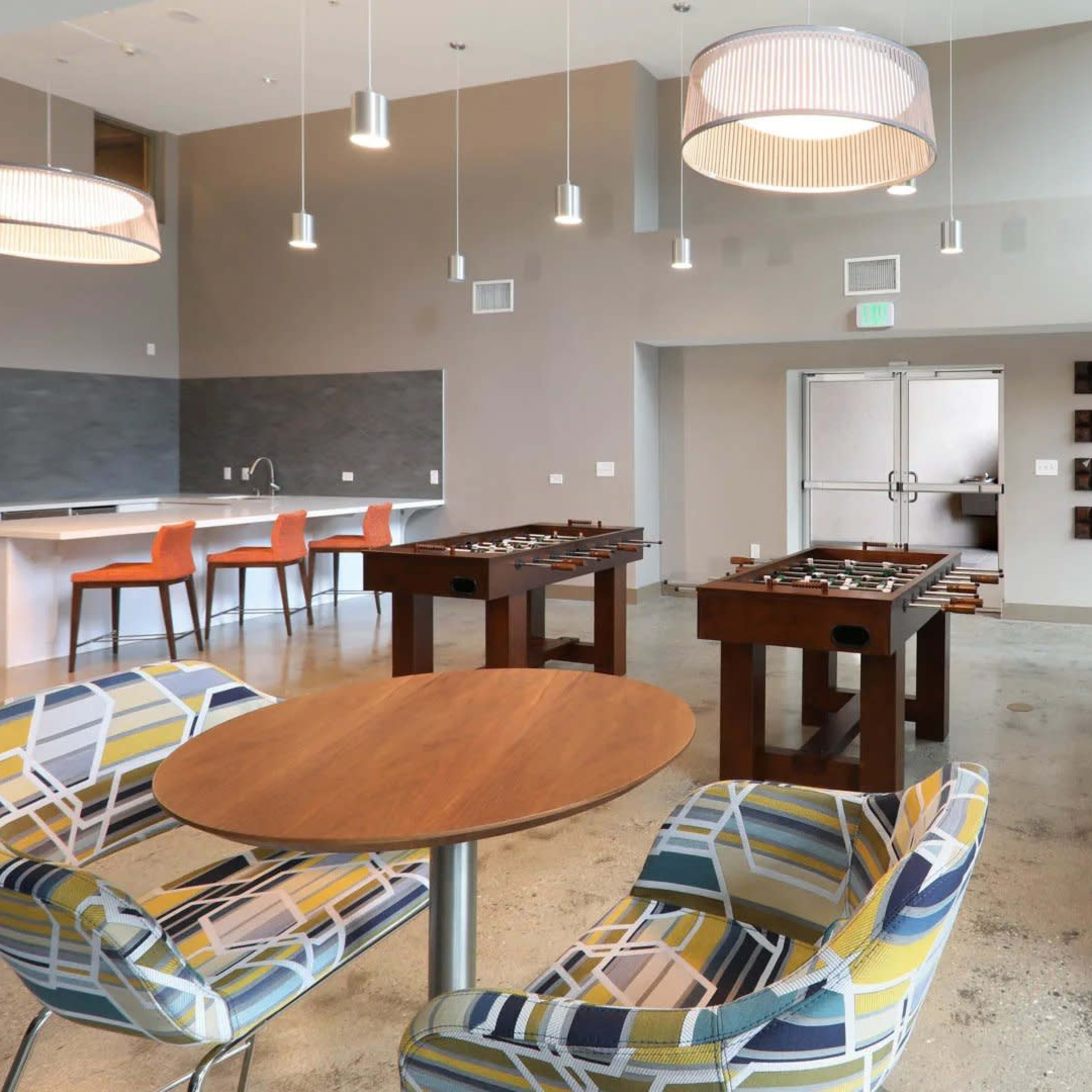 A modern recreational room with a dining table, colorful chairs, foosball tables, and a shuffleboard table, all set in a well-lit space.