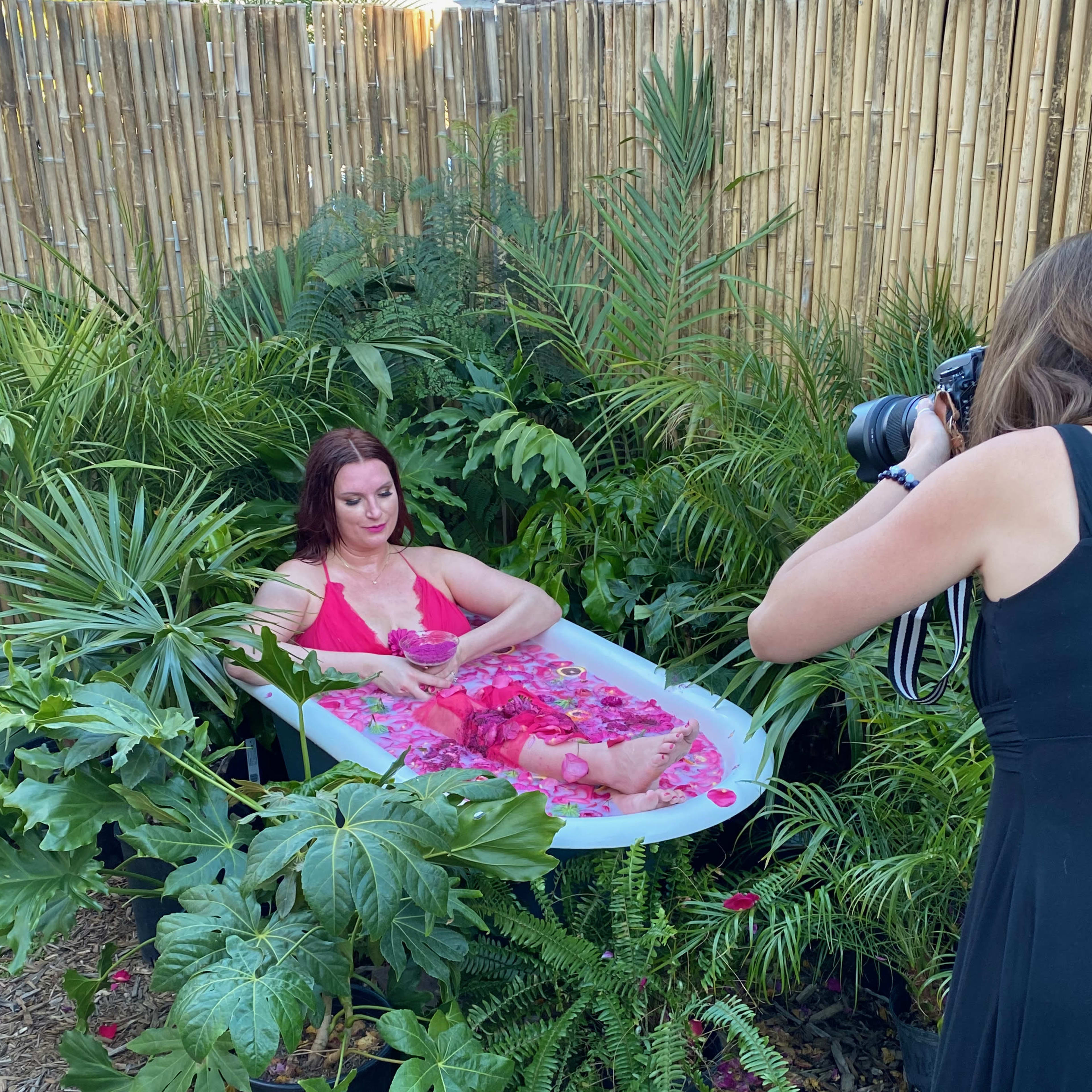 A woman in a bath surrounded by tropical plants poses for a photograph, while another woman in a black dress captures the moment with a camera.