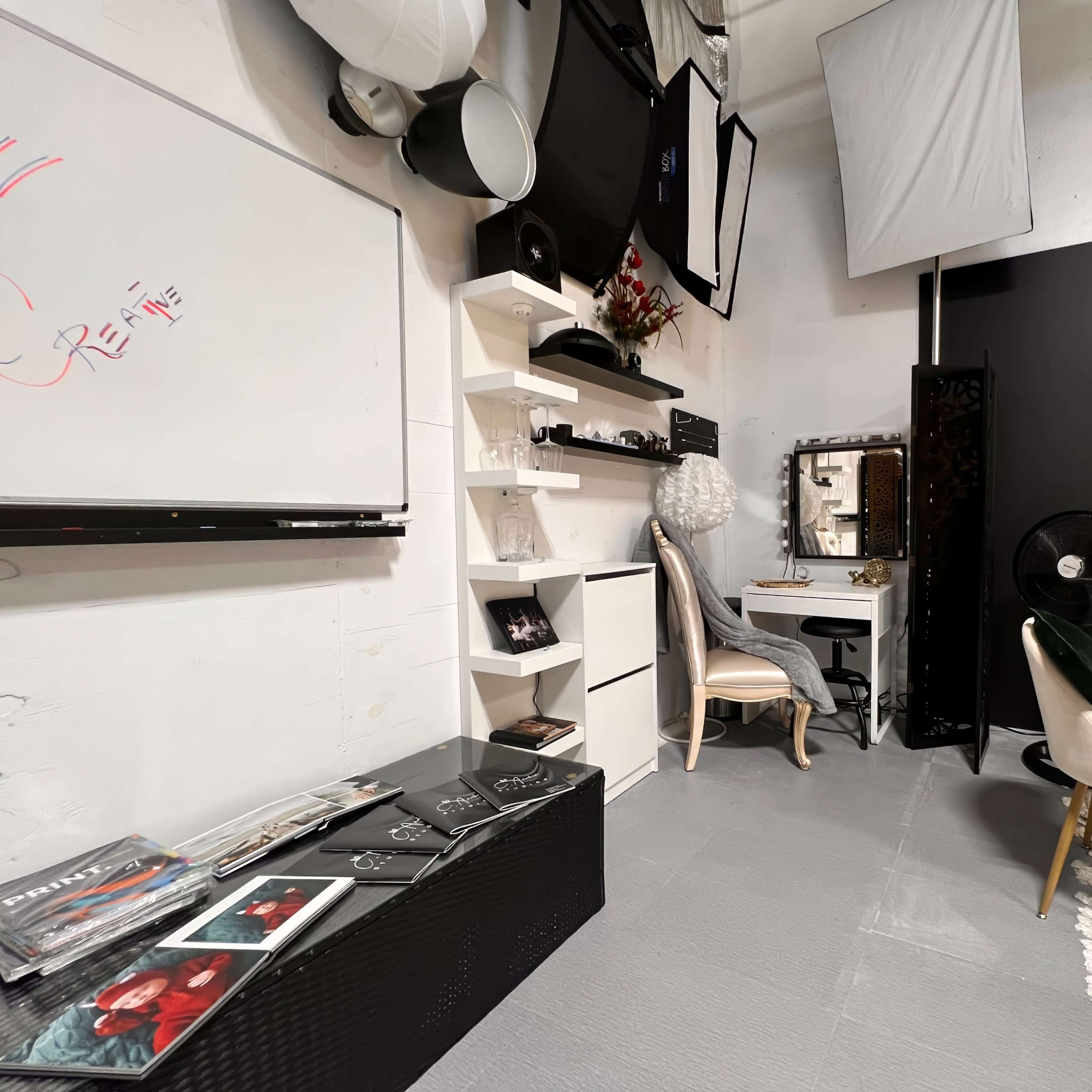 A neatly organized studio space featuring a whiteboard, shelves with decorative items, a dressing table, seating, and a backdrop against a wall.