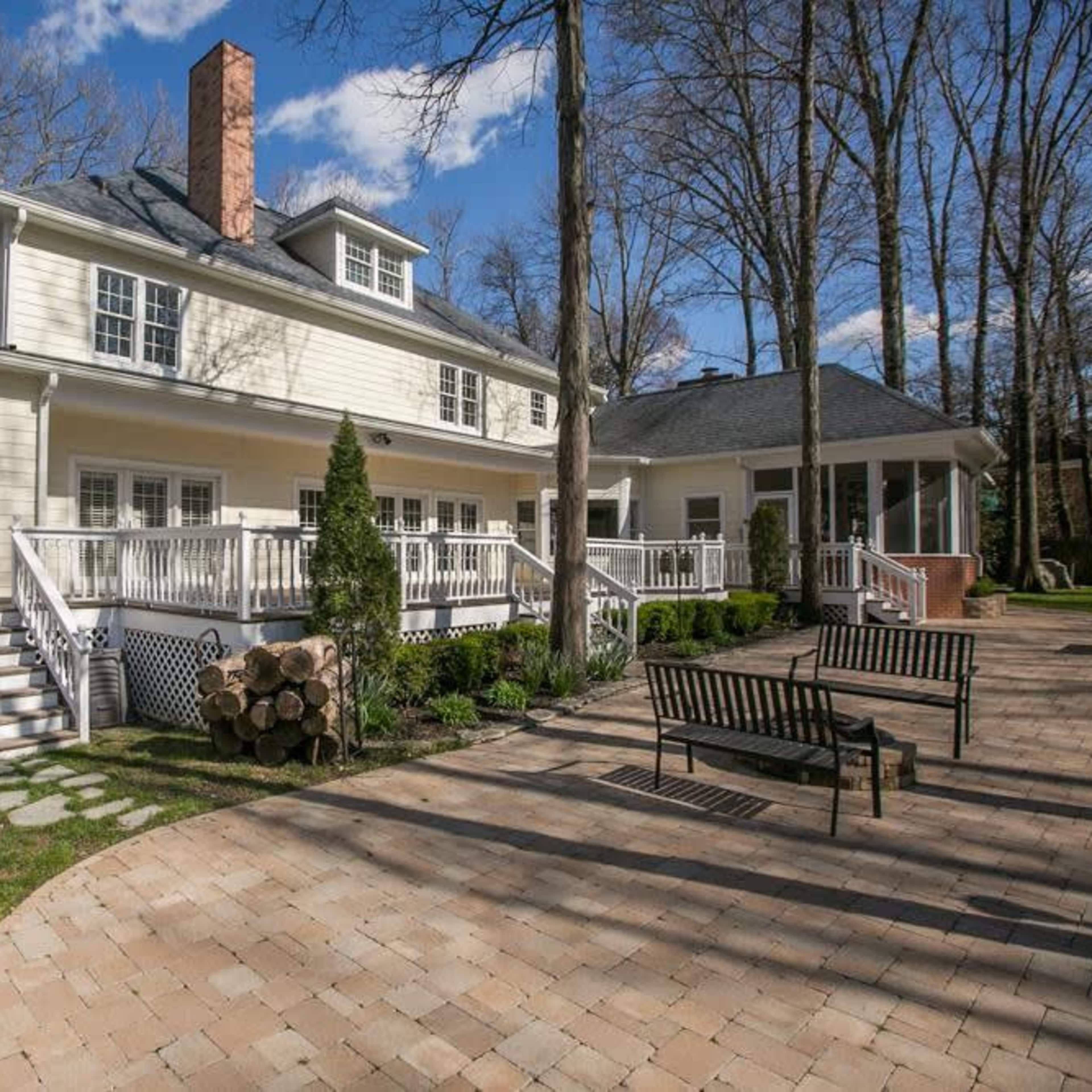 A large, light-colored house with a sloped roof, a brick chimney, a spacious deck, and a paved patio area surrounded by trees.