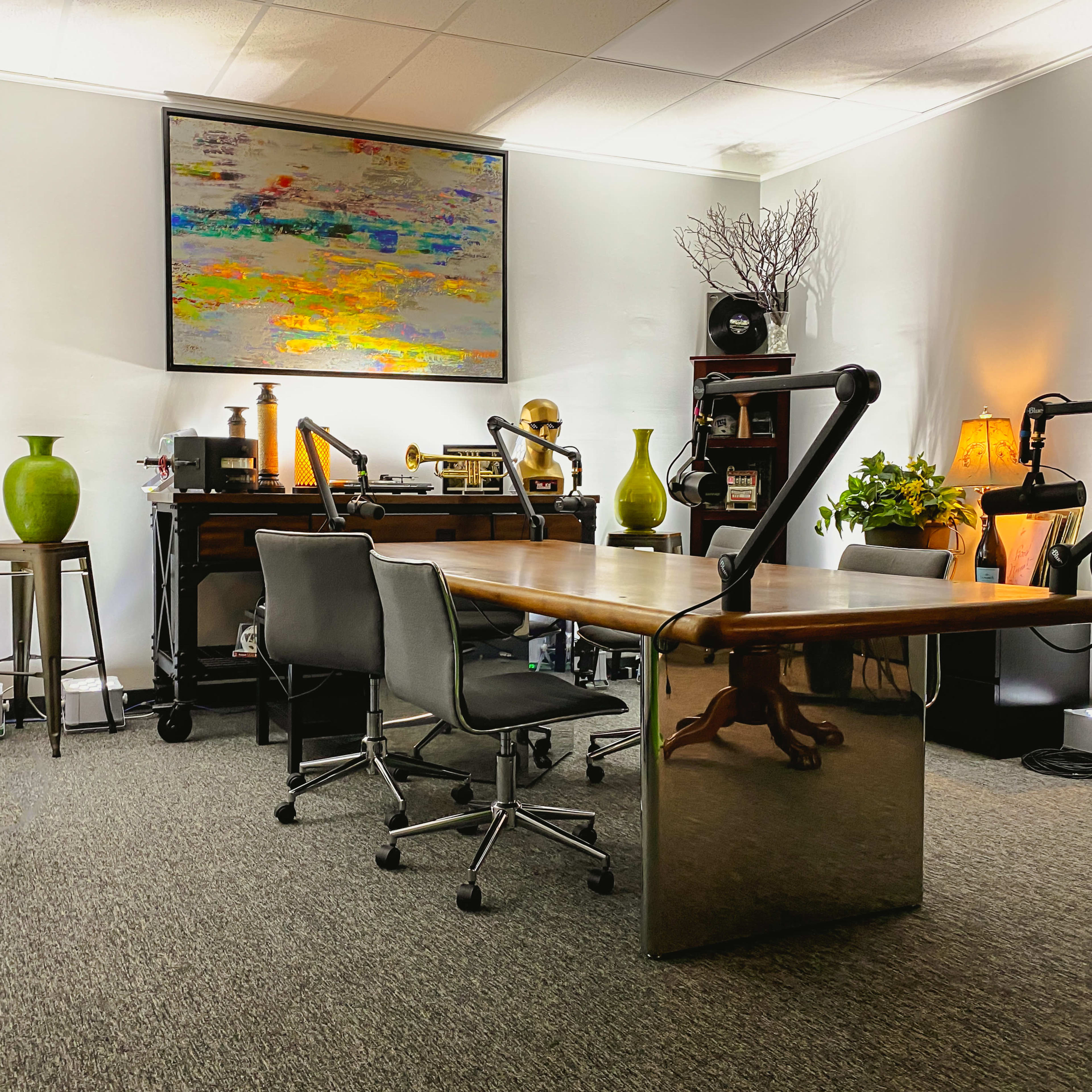 A modern office space features a large wooden table with two chairs, surrounded by plants, decor, and a colorful painting on the wall.