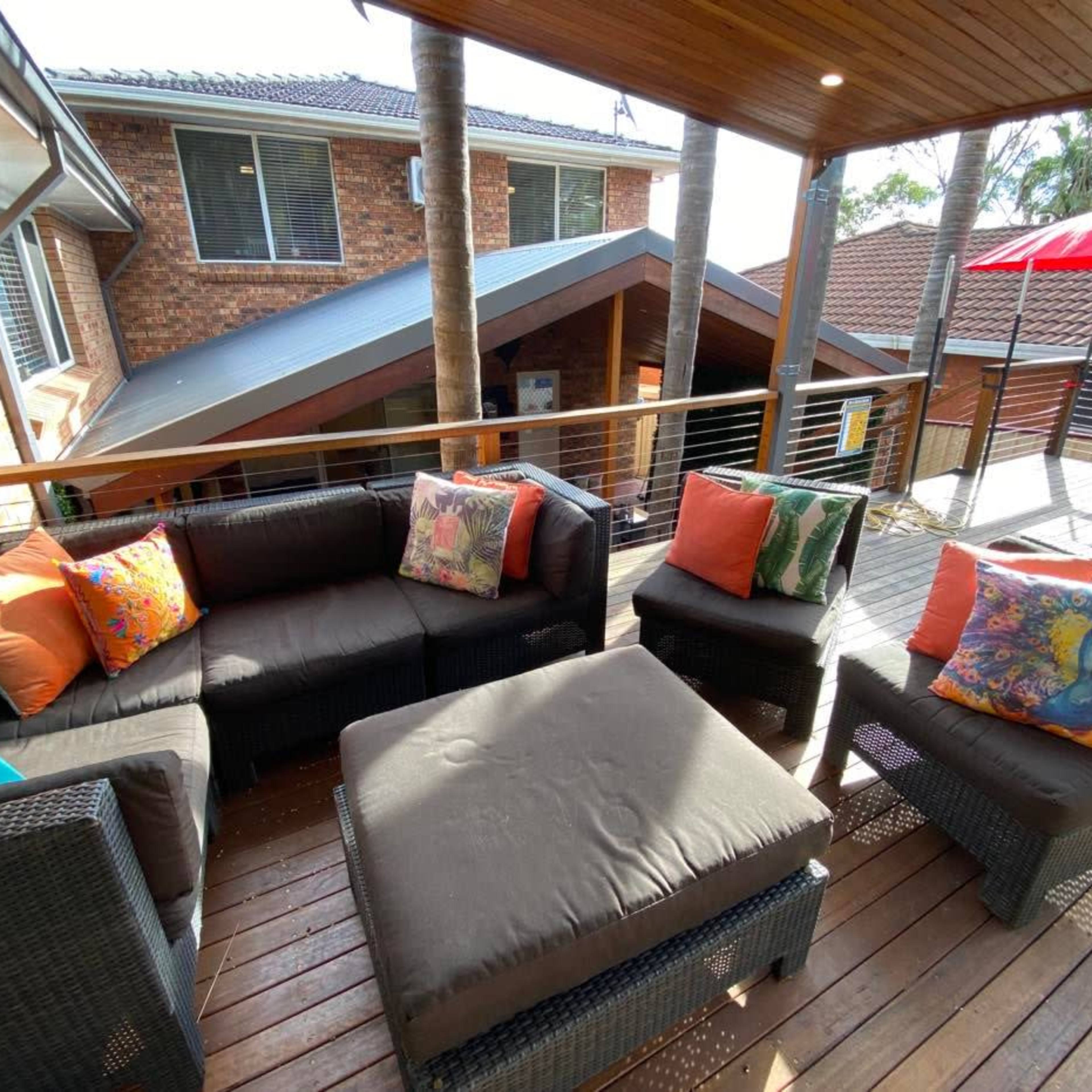 A covered outdoor seating area with a wicker couch, colorful pillows, and a square coffee table on a wooden deck.