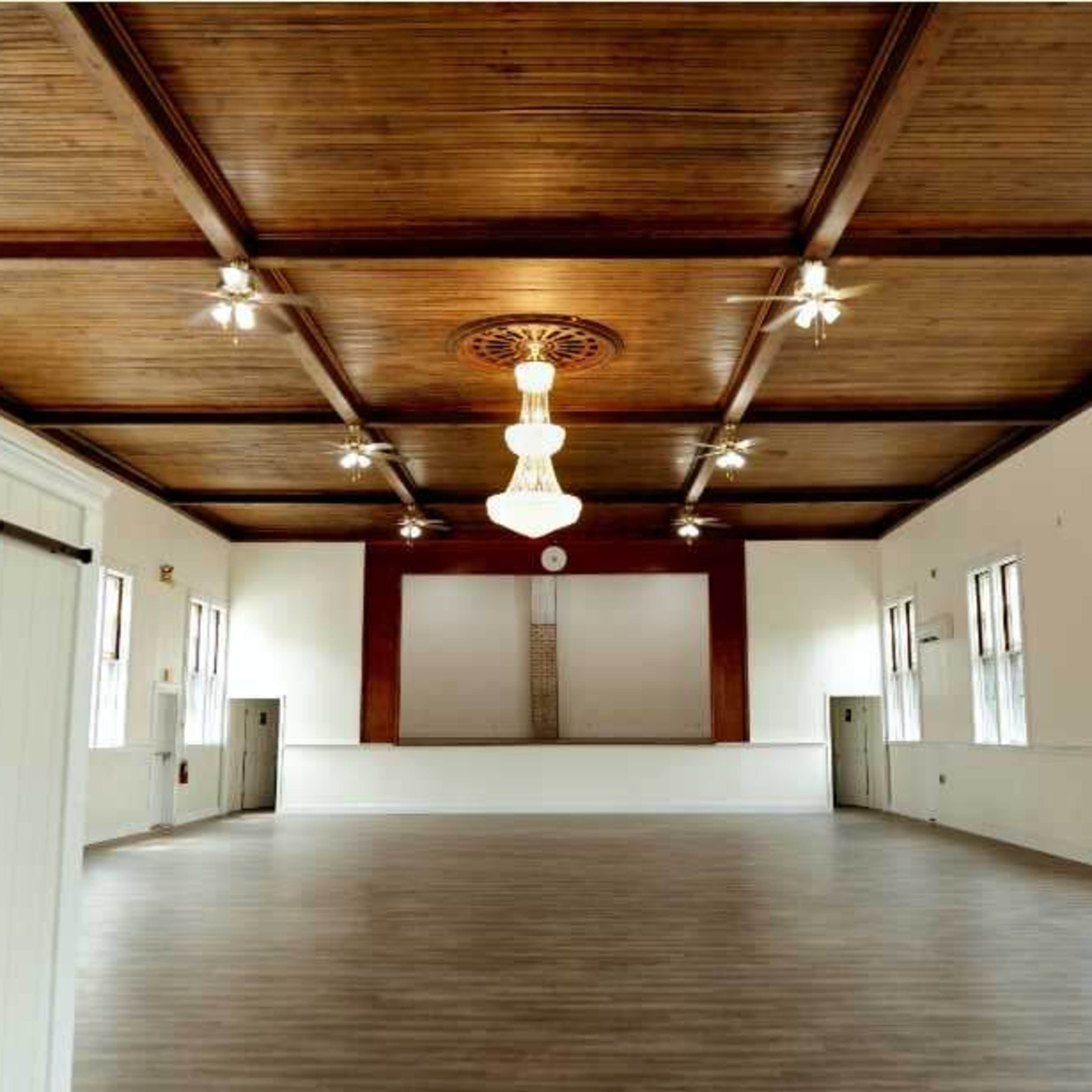 An empty indoor hall with a wooden ceiling, chandeliers, and a stage at one end.