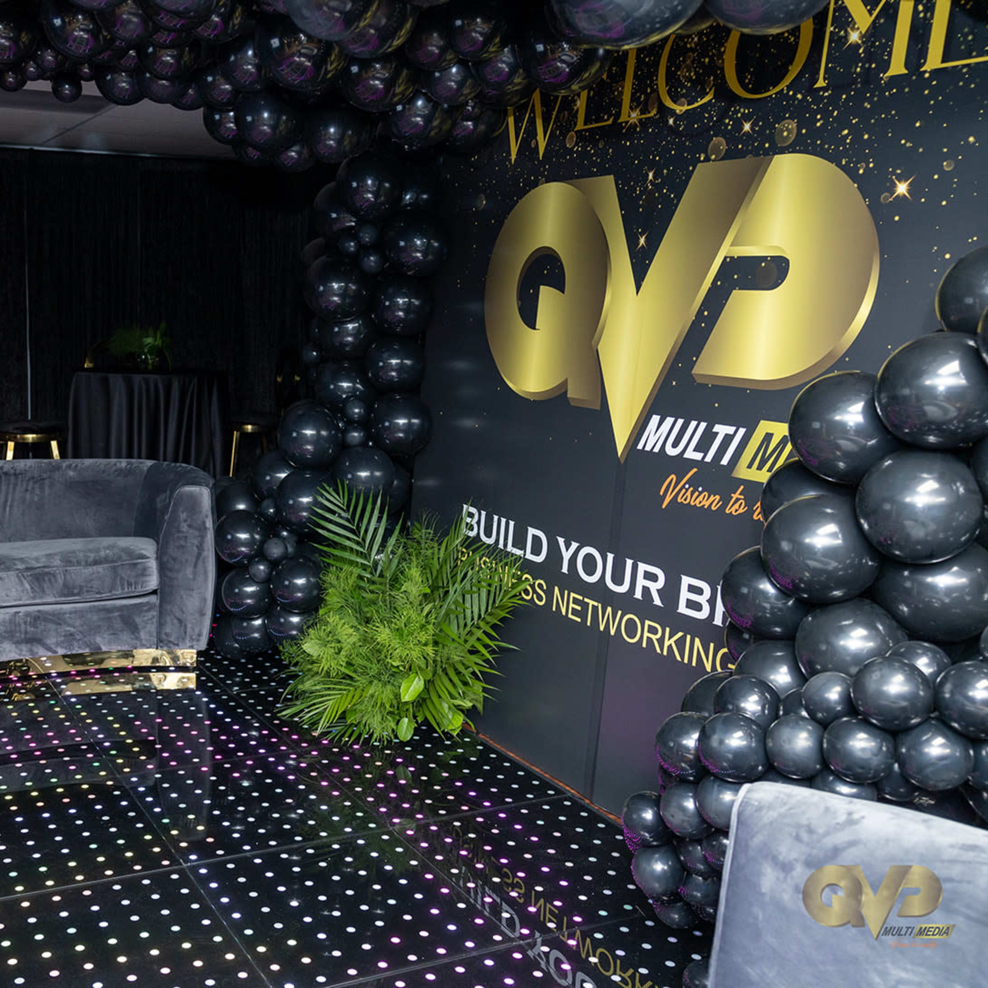 A dimly lit event space decorated with black and gold balloons, featuring a plush gray sofa, small tables, and a branded backdrop promoting a business networking event.