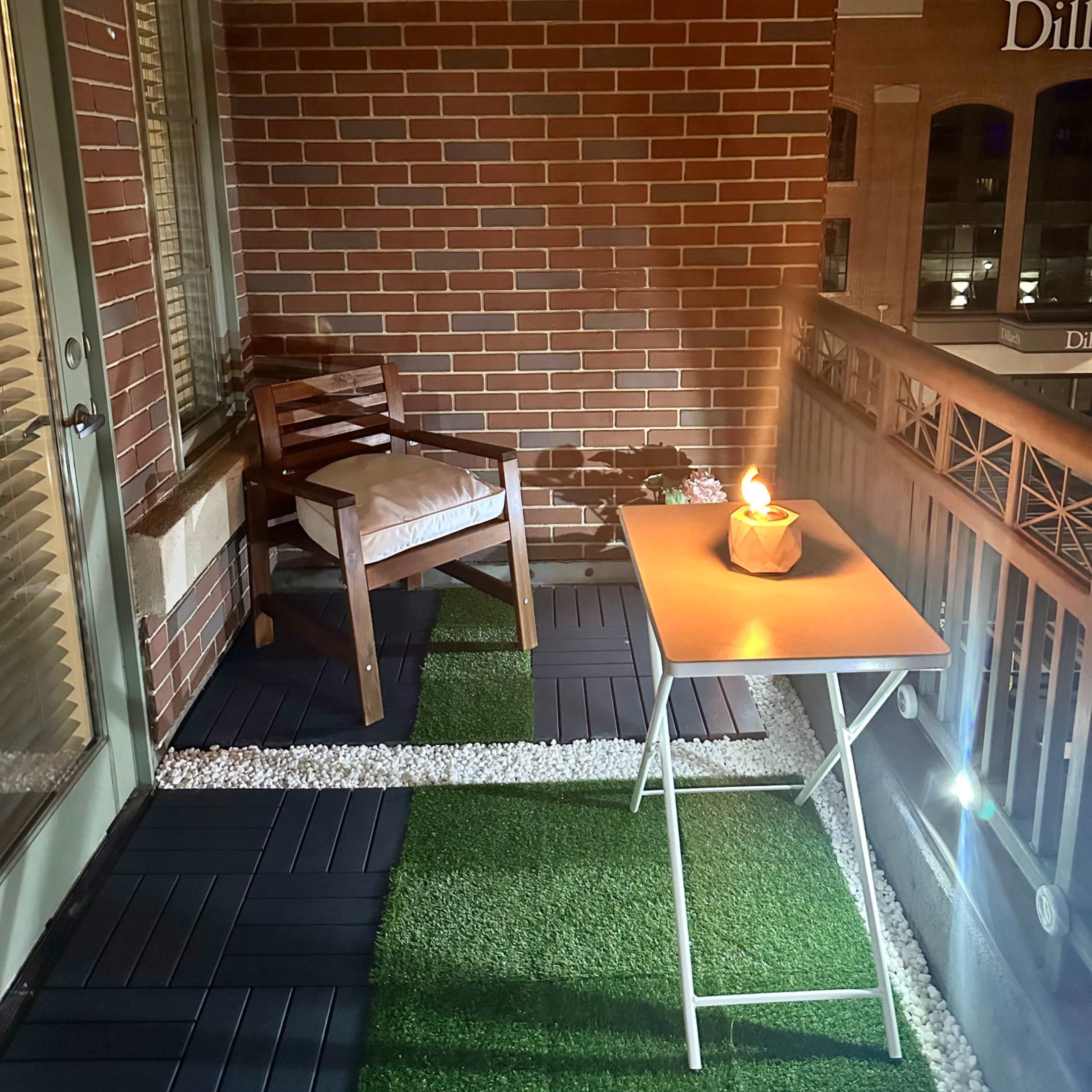 A small balcony with a wooden chair and a folding table, both illuminated by a candle, set against a backdrop of brick walls and a cityscape.