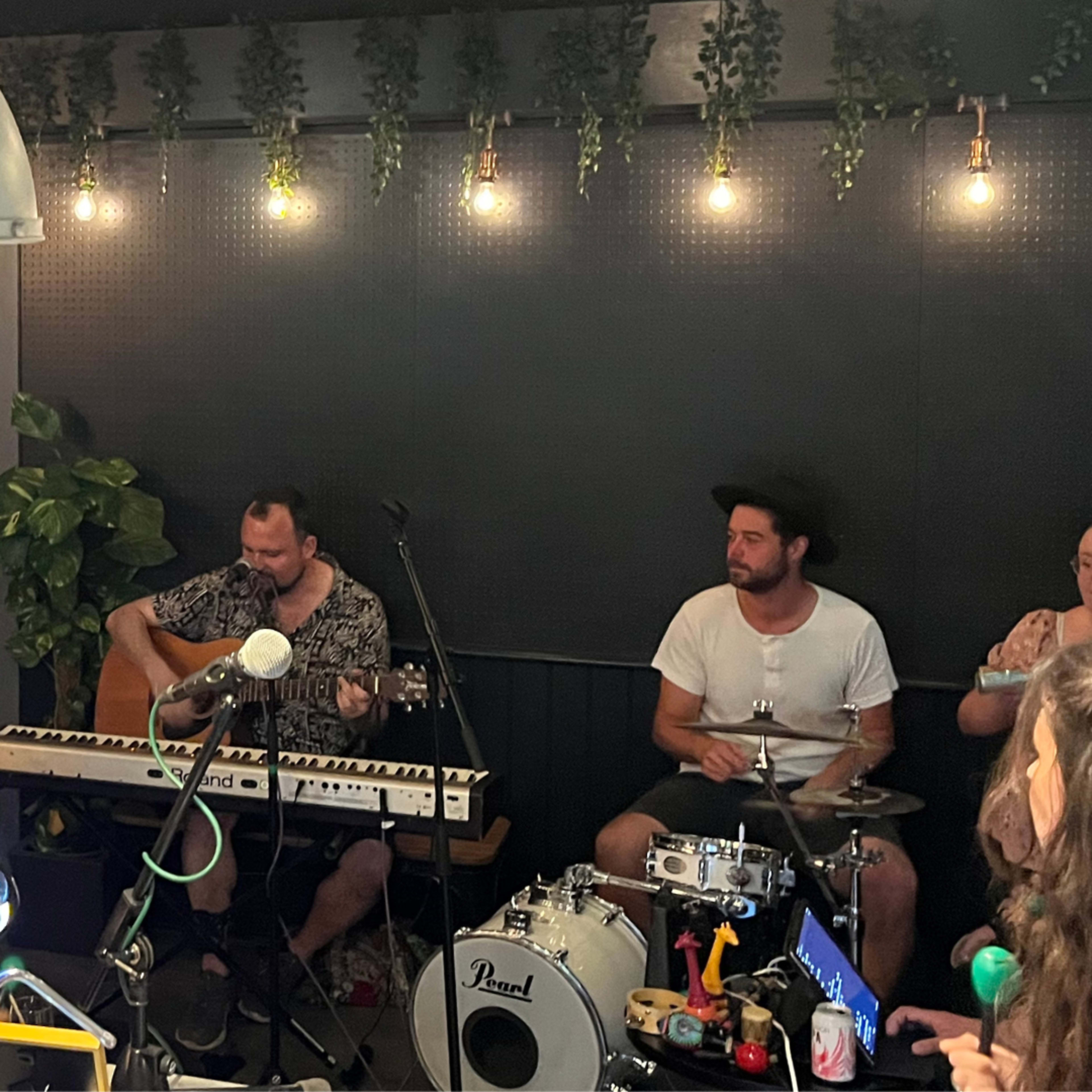Two musicians perform in a dimly lit room, one playing guitar and the other drumming, surrounded by plants and string lights.
