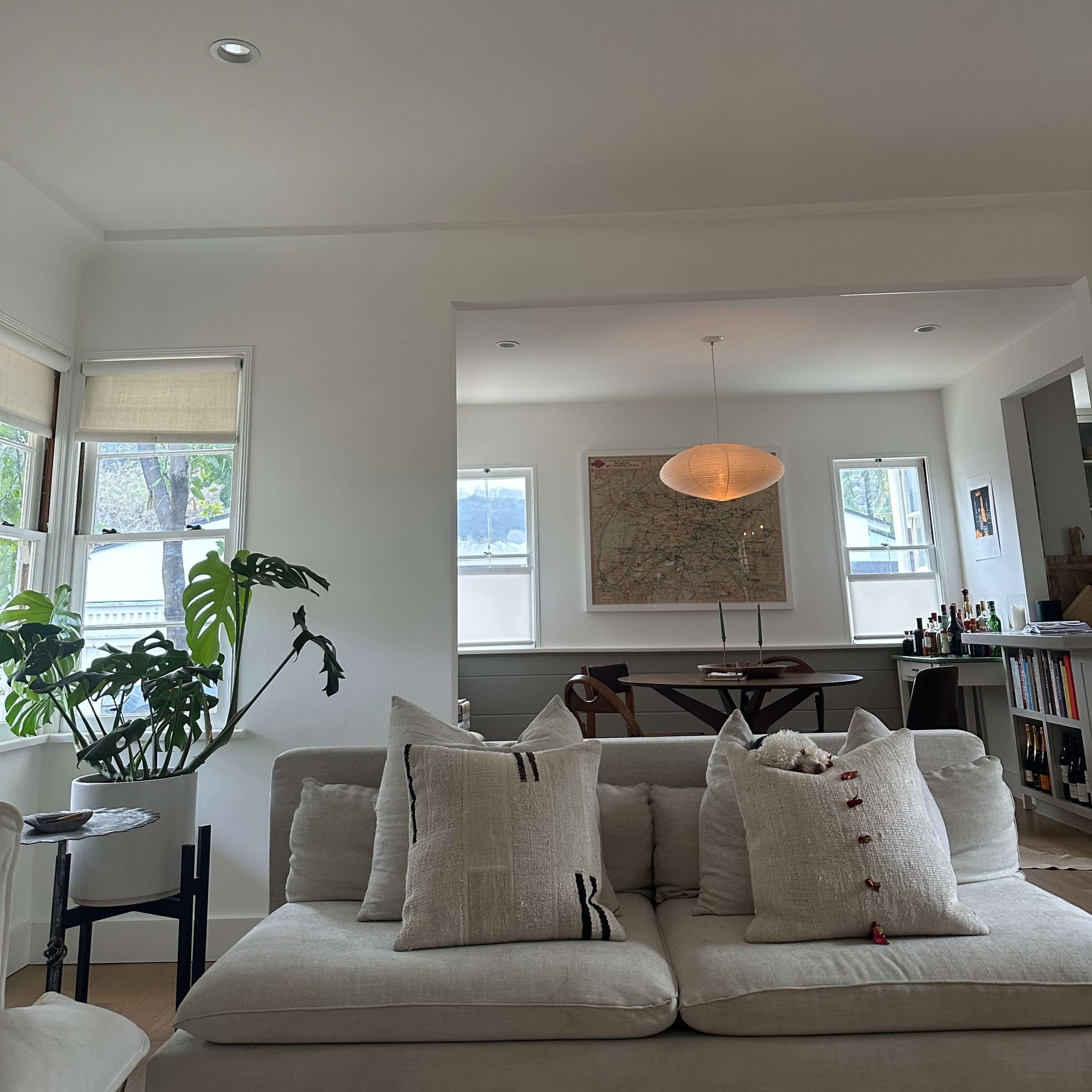 A living room with a light-colored sofa adorned with decorative pillows, a dining area in the background, and large windows allowing natural light to enter.