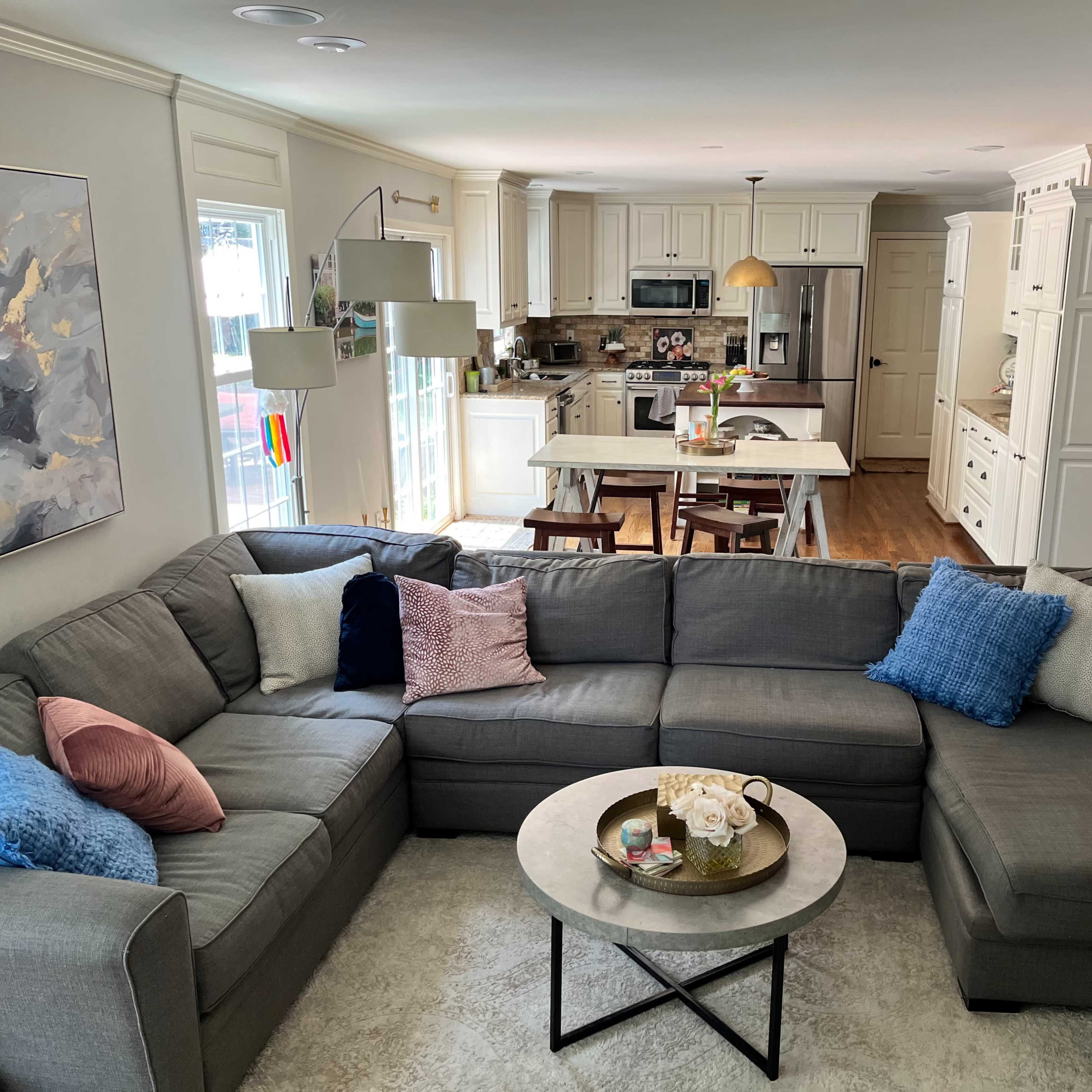 A spacious living room features a large gray sectional sofa with colorful throw pillows and a circular coffee table, overlooking an open kitchen with white cabinetry.