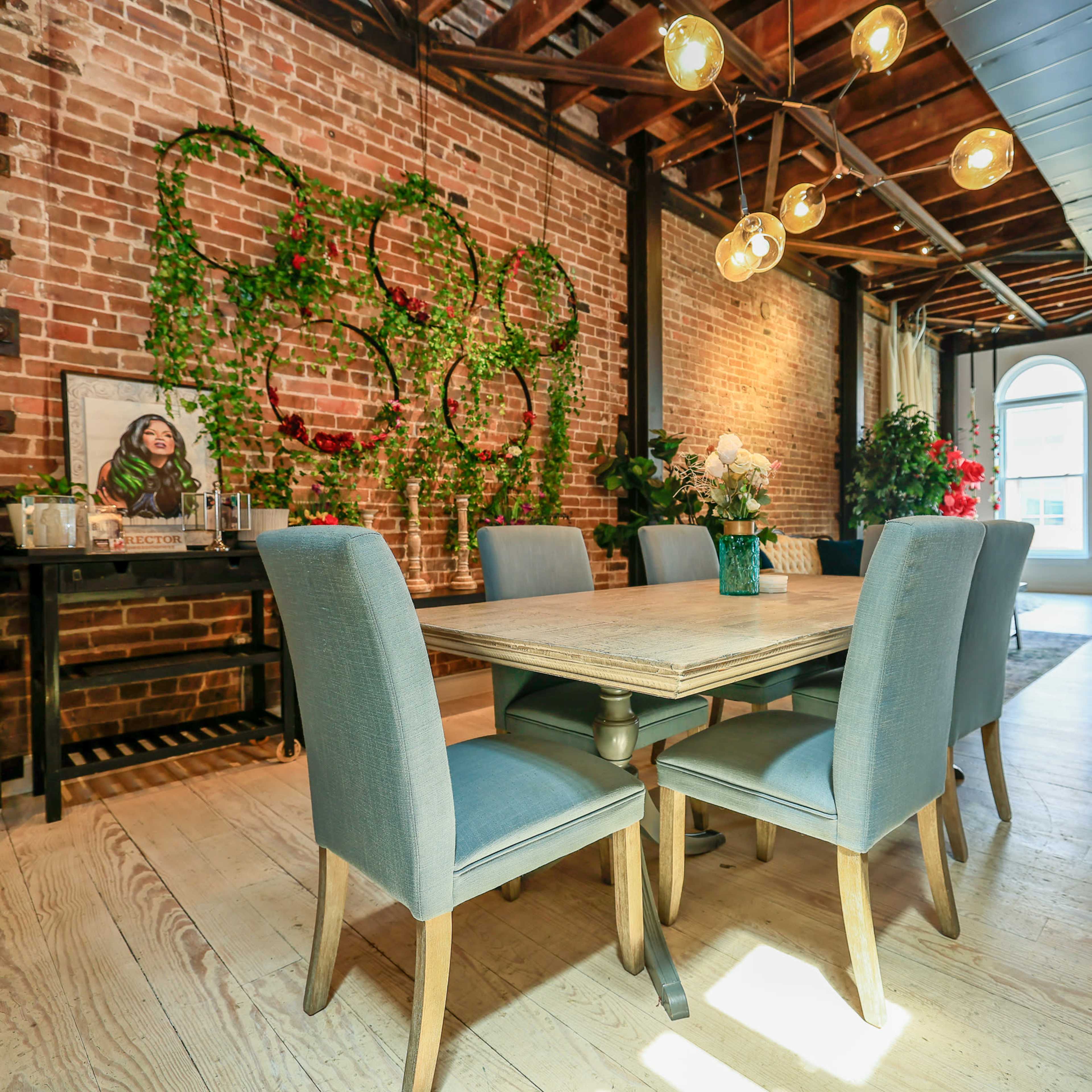 A dining area features a wooden table surrounded by upholstered chairs, with a brick wall adorned with greenery and pendant lights above.
