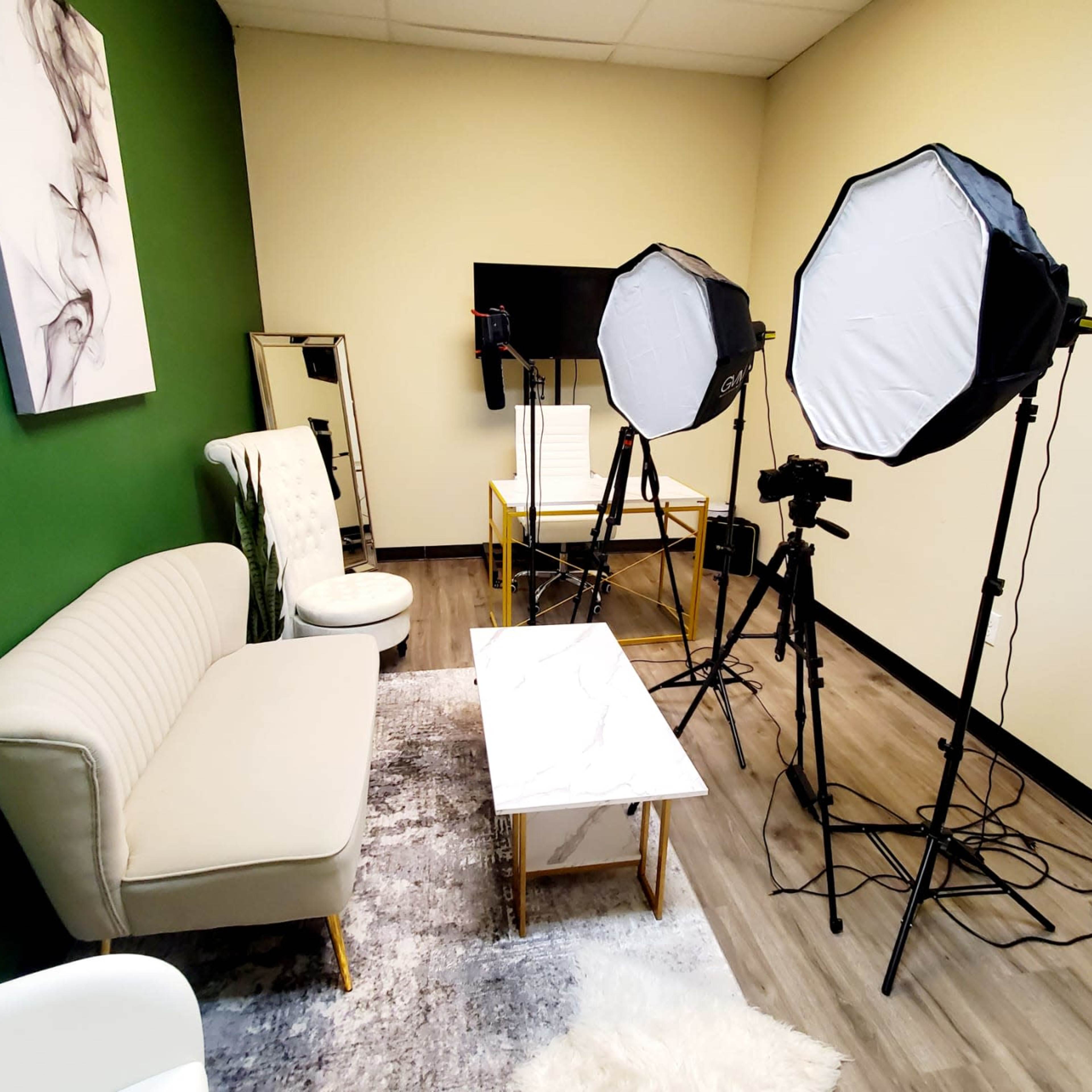 A small studio setup with two softbox lights, camera equipment, a minimalist sofa, a coffee table, and a green accent wall.