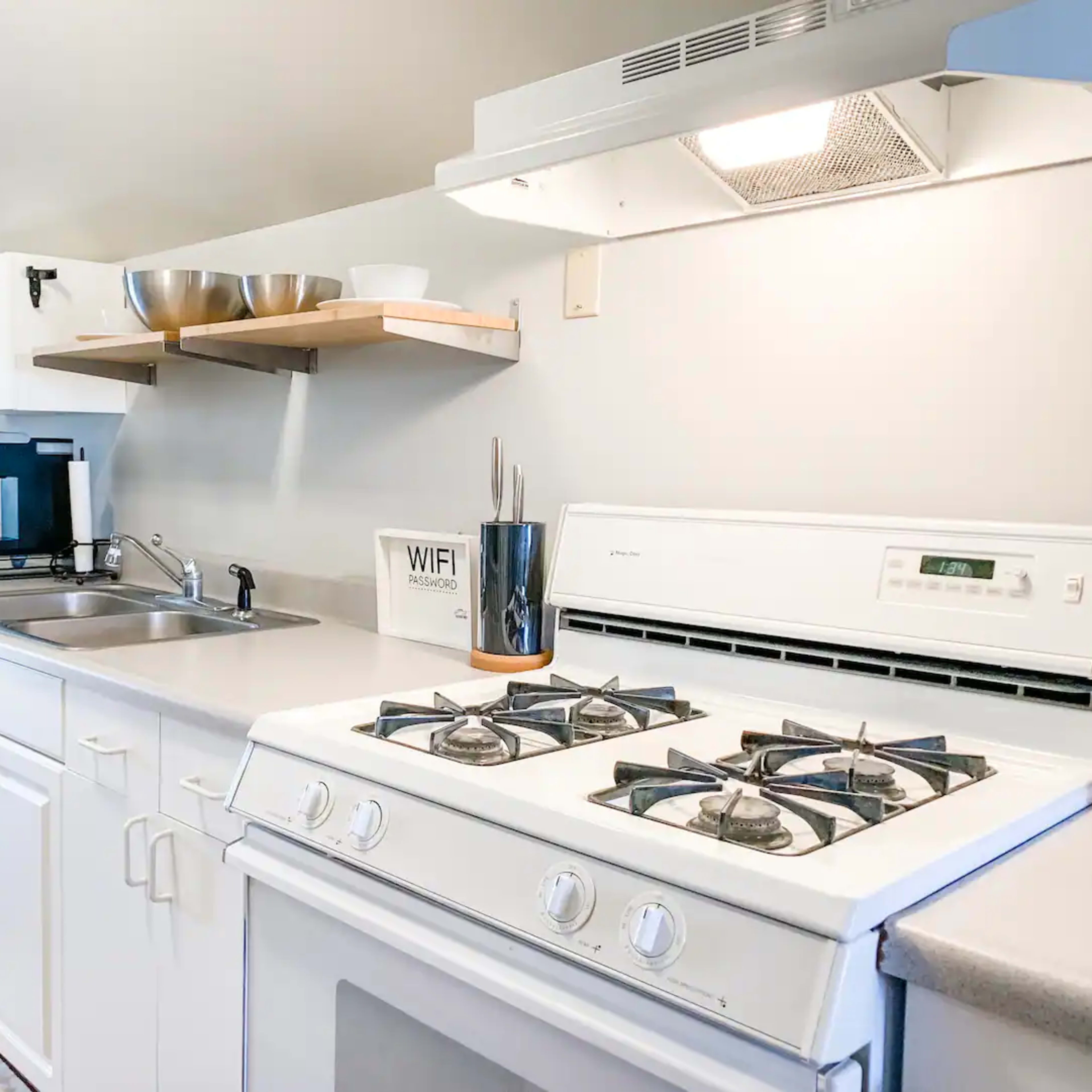 A modern kitchen with white cabinets, a sink, a stove, and a coffee maker on the counter, alongside open shelving with bowls and a decorative sign.