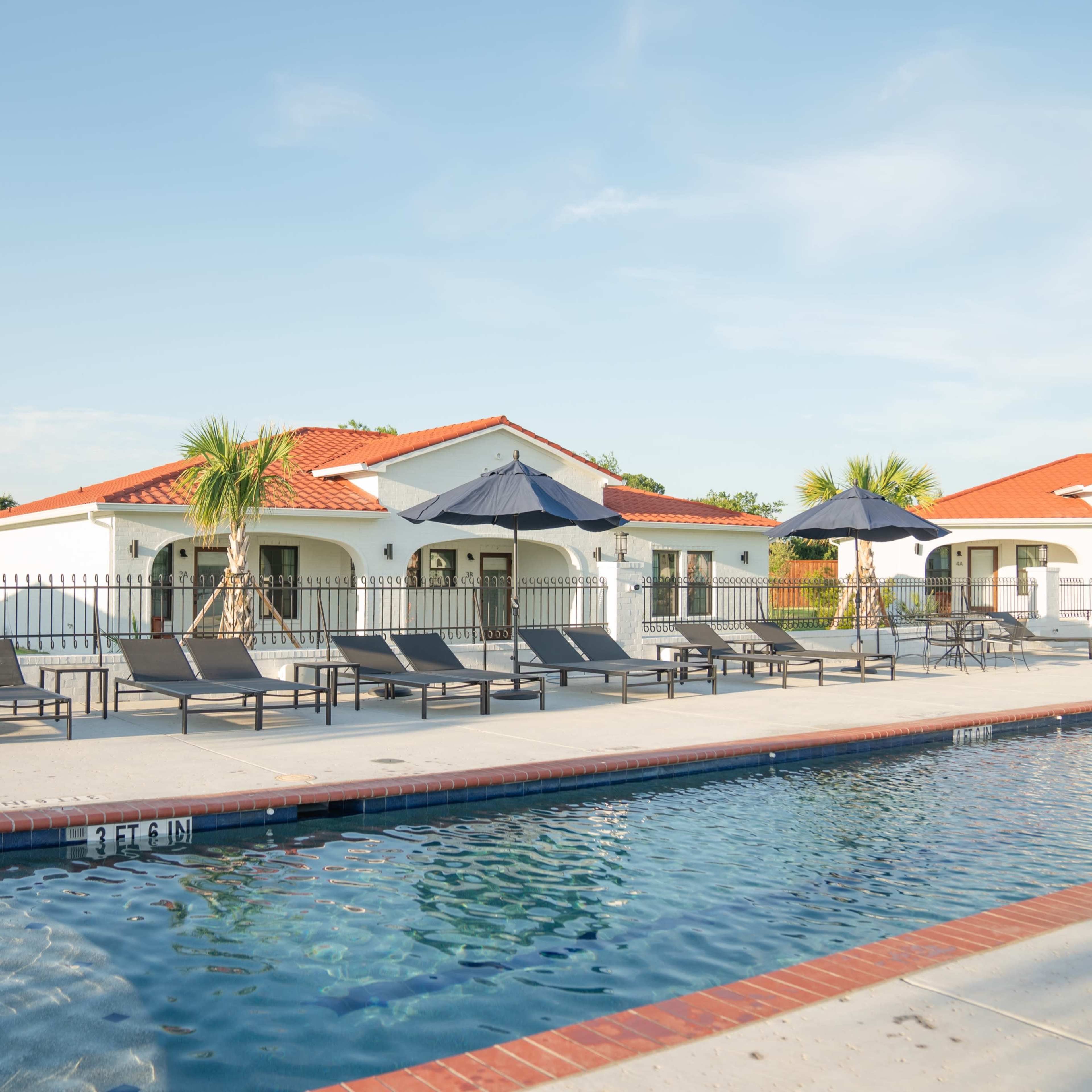 A swimming pool with lounge chairs and umbrellas is situated beside a white building with a red-tiled roof.
