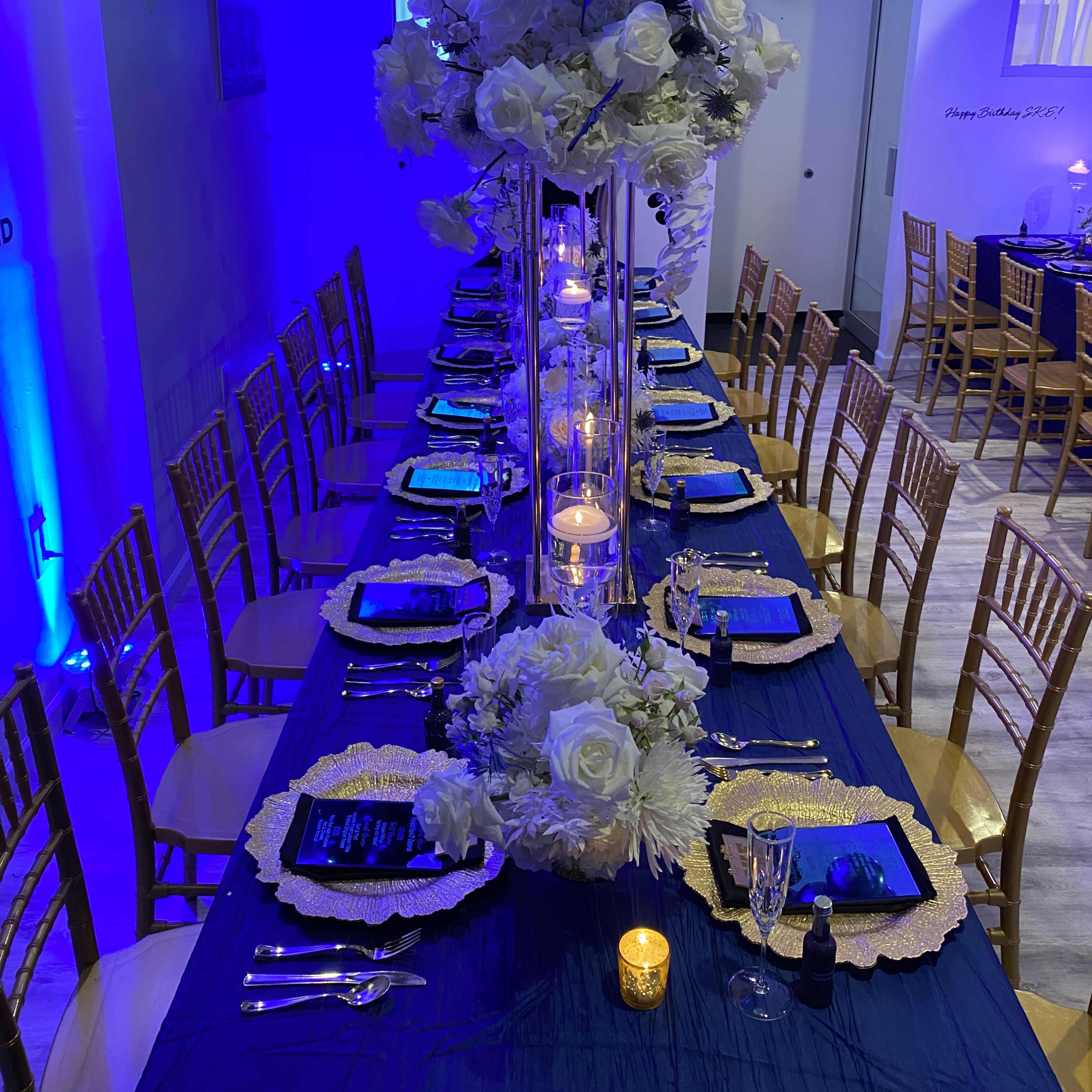 A long banquet table is elegantly set with gold-accented plates, surrounded by wooden chairs, and adorned with white flowers and a cluster of blue and black balloons overhead.