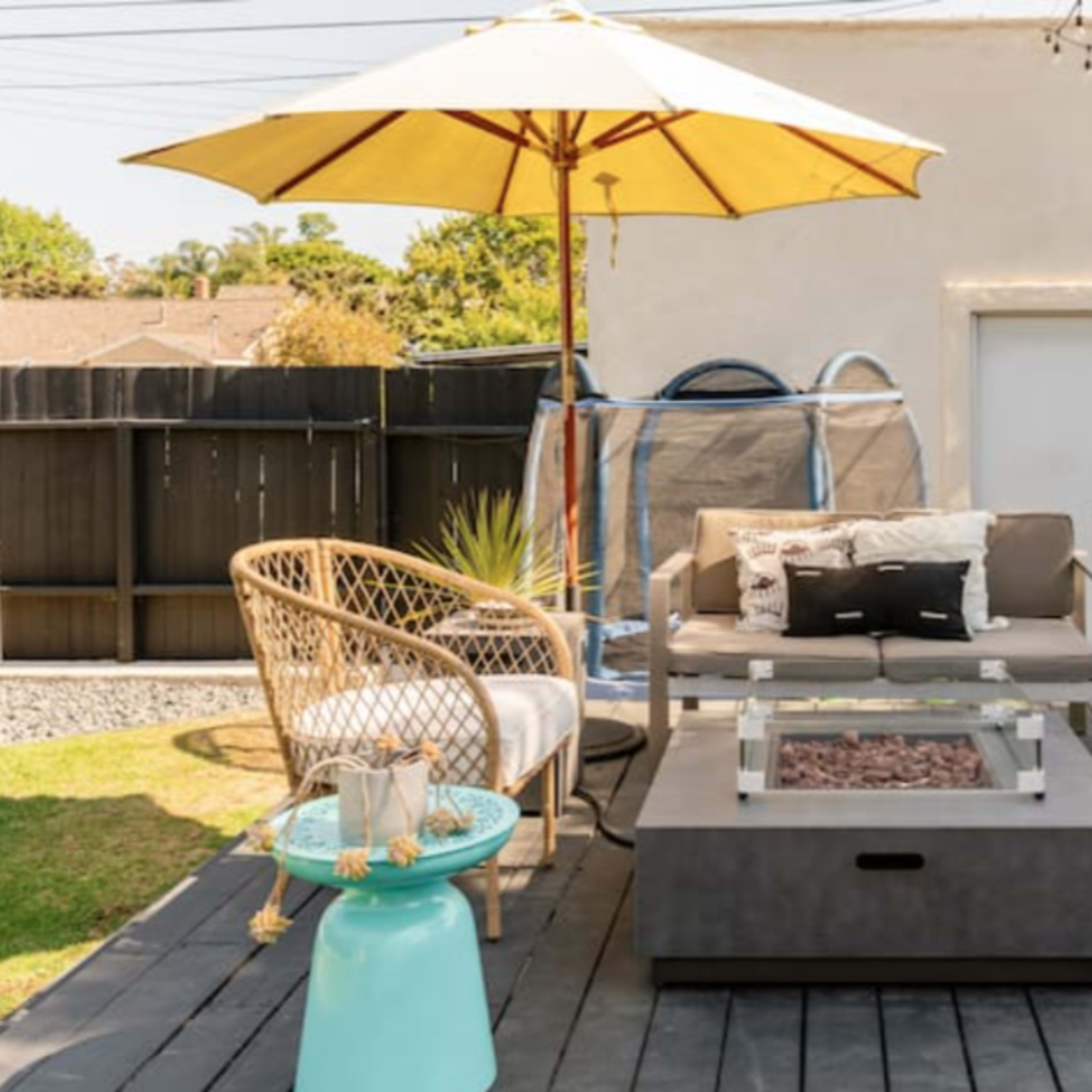 A patio area featuring a couch with an umbrella, two woven chairs, a coffee table with a fire pit, and a trampoline in the background.