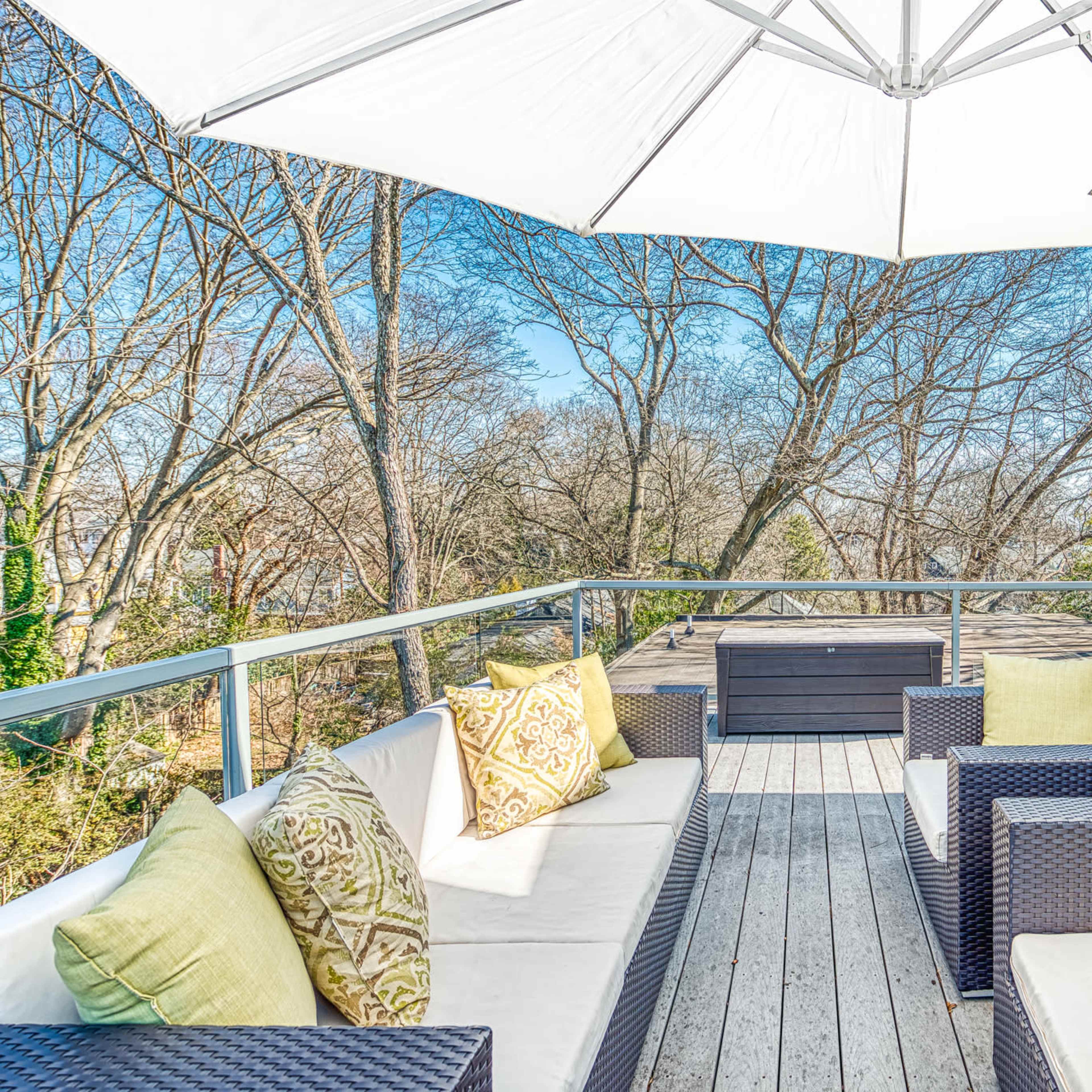 A spacious outdoor terrace features woven furniture, pale cushions, and a large umbrella overlooking a landscape of bare trees.