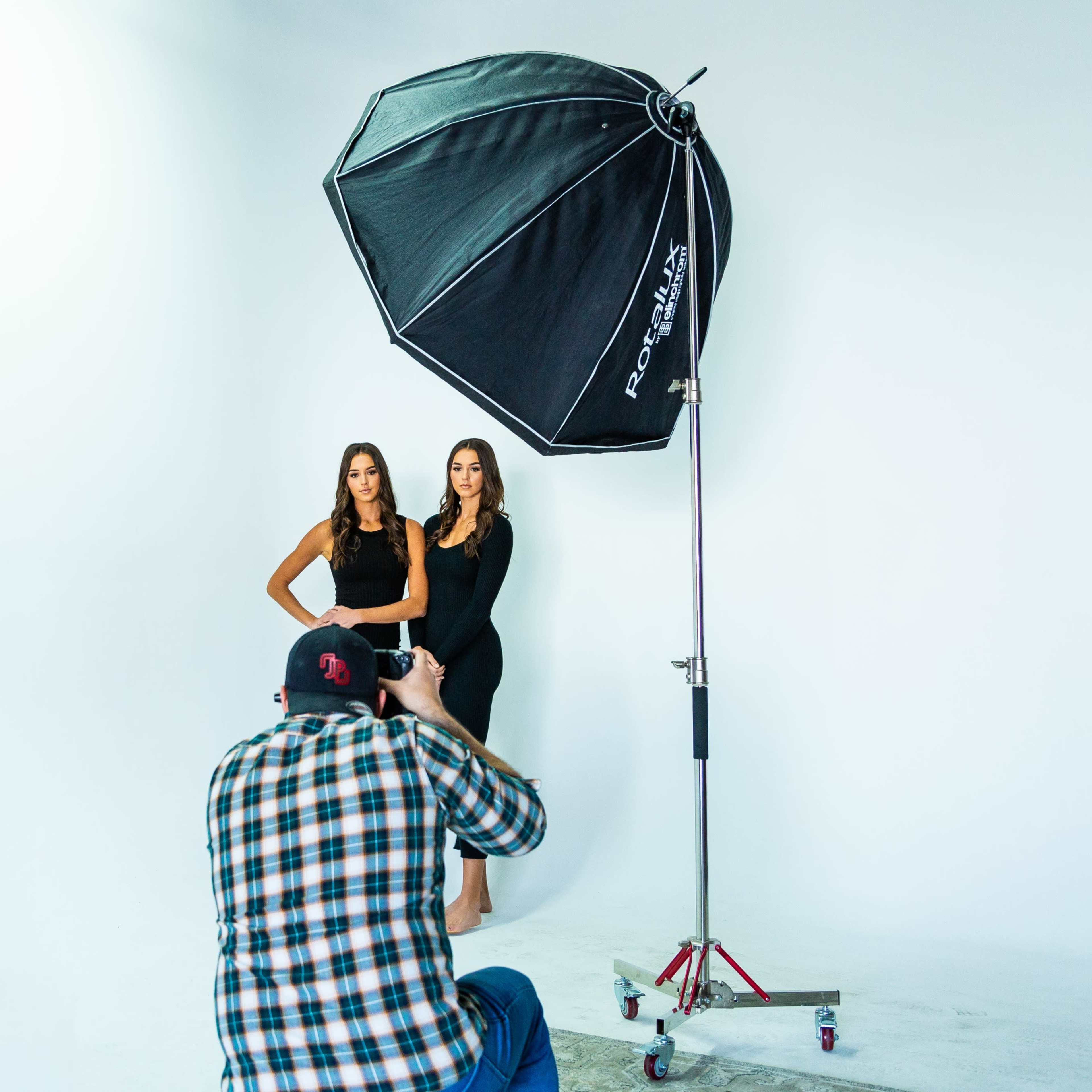 A photographer captures an image of two models posing in a studio with a large umbrella light above them.