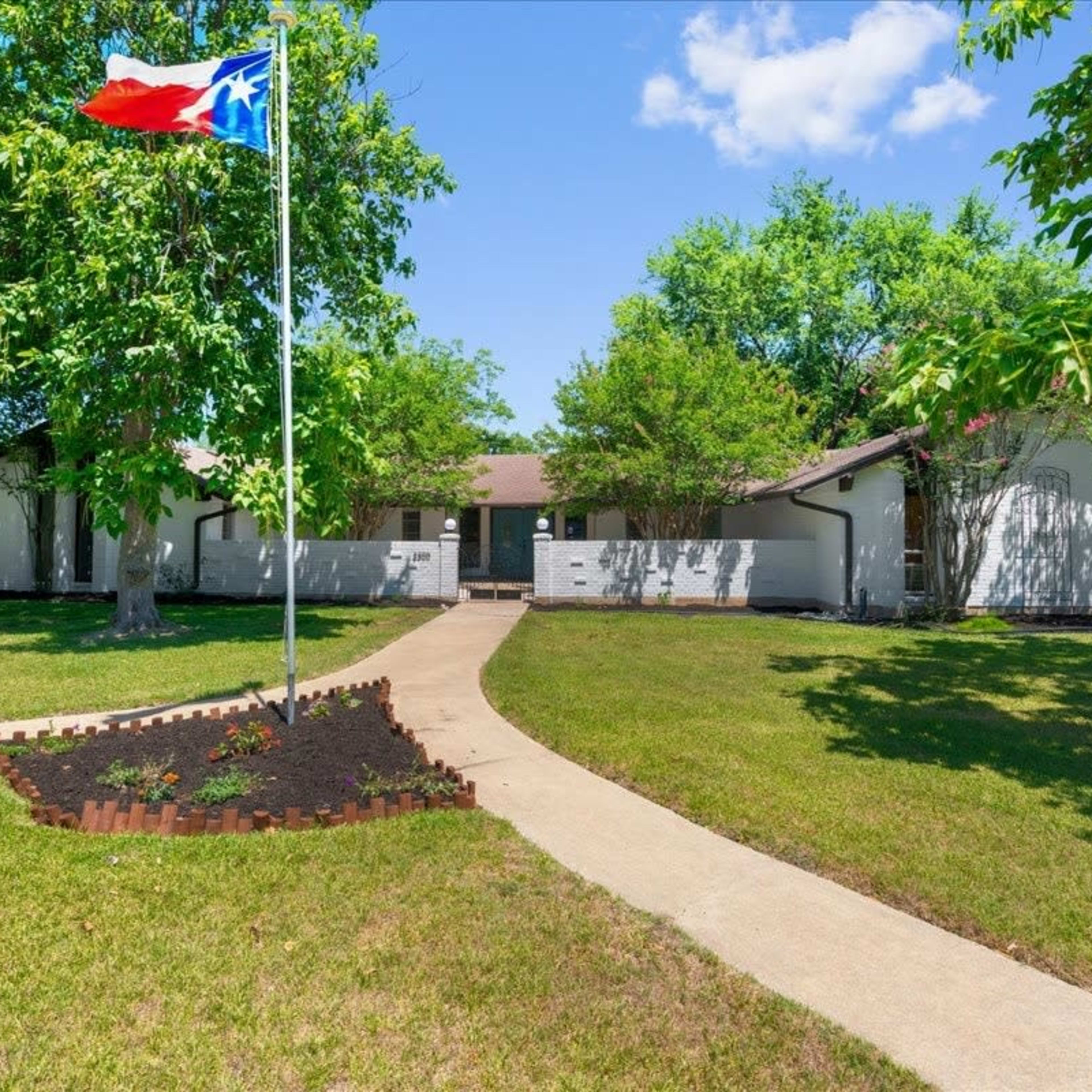 A single-story house with a Texas flag on a flagpole in the front yard, surrounded by green grass and trees.