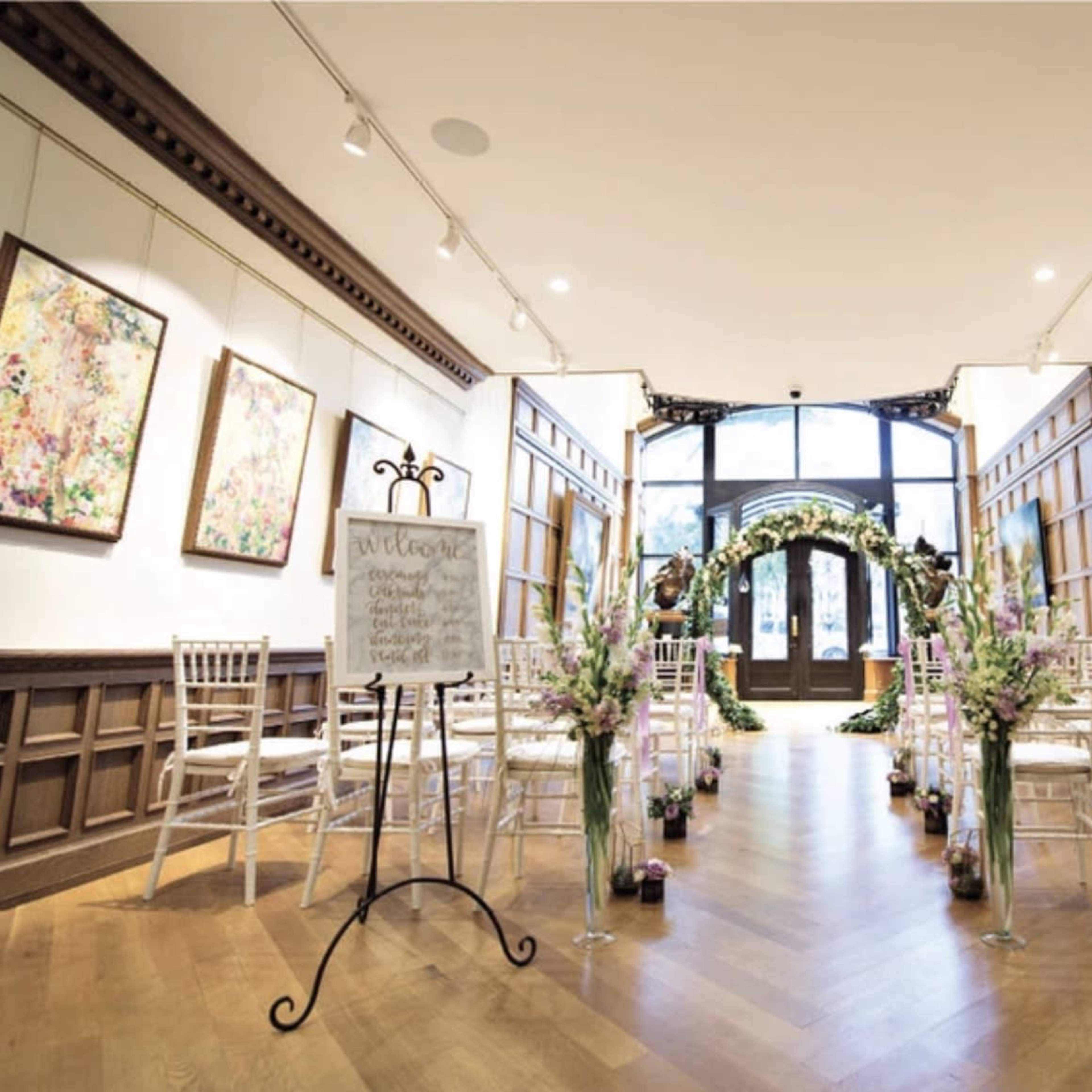 A wedding ceremony setup features rows of white chairs facing an ornate arch adorned with greenery and flowers in a tastefully decorated gallery.