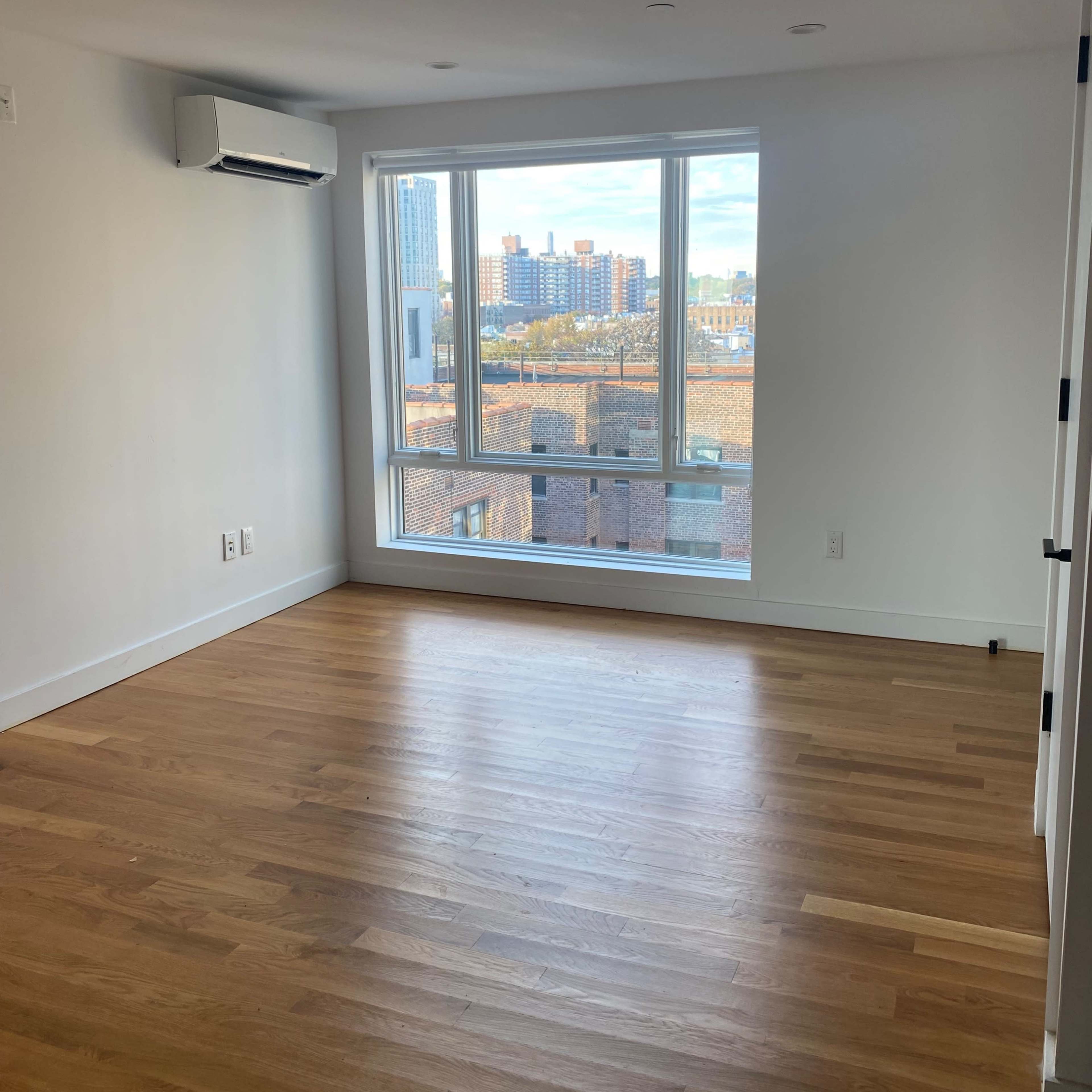 A vacant room with hardwood flooring and a large window overlooking a cityscape.