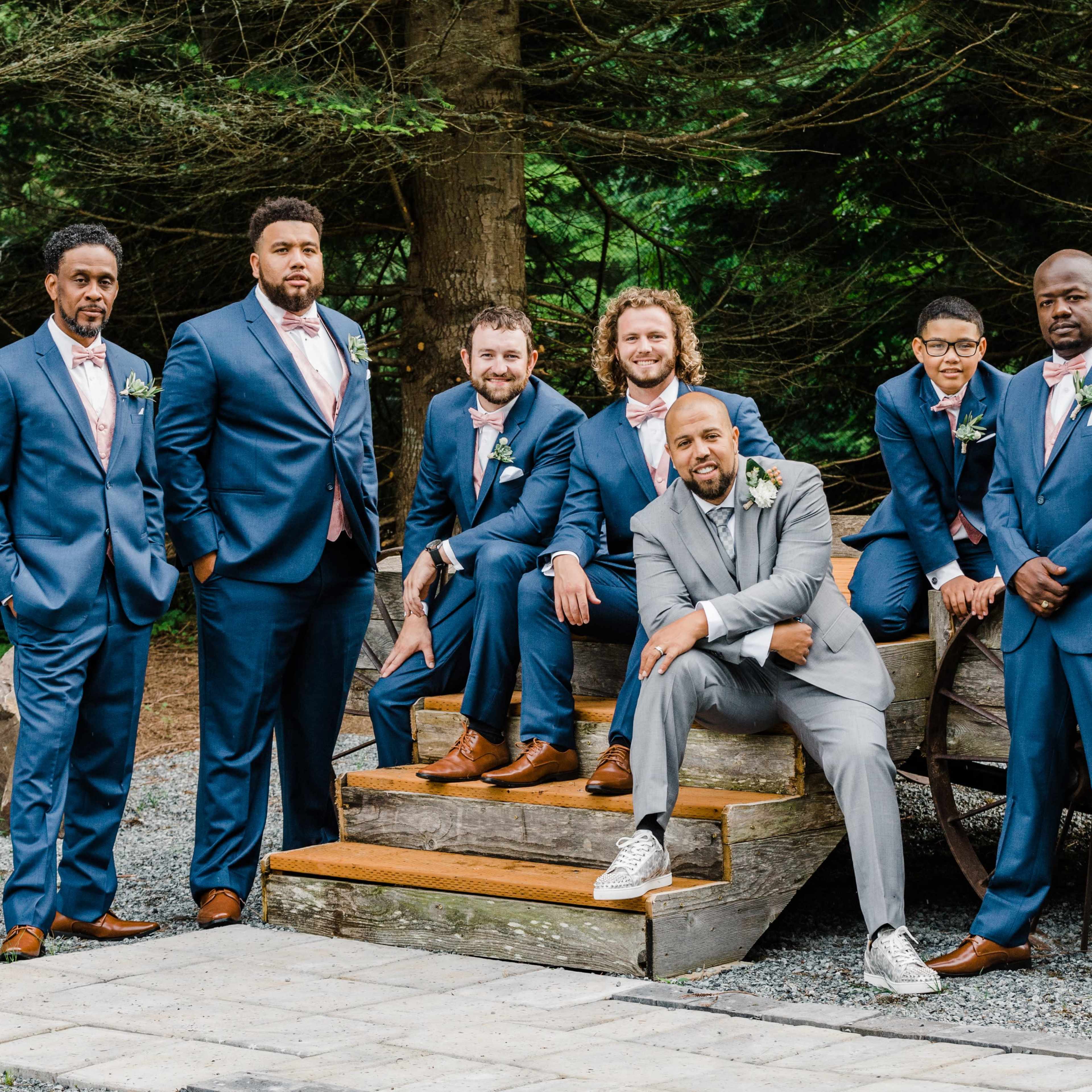 A group of seven men in formal attire poses on stone steps in a wooded area.