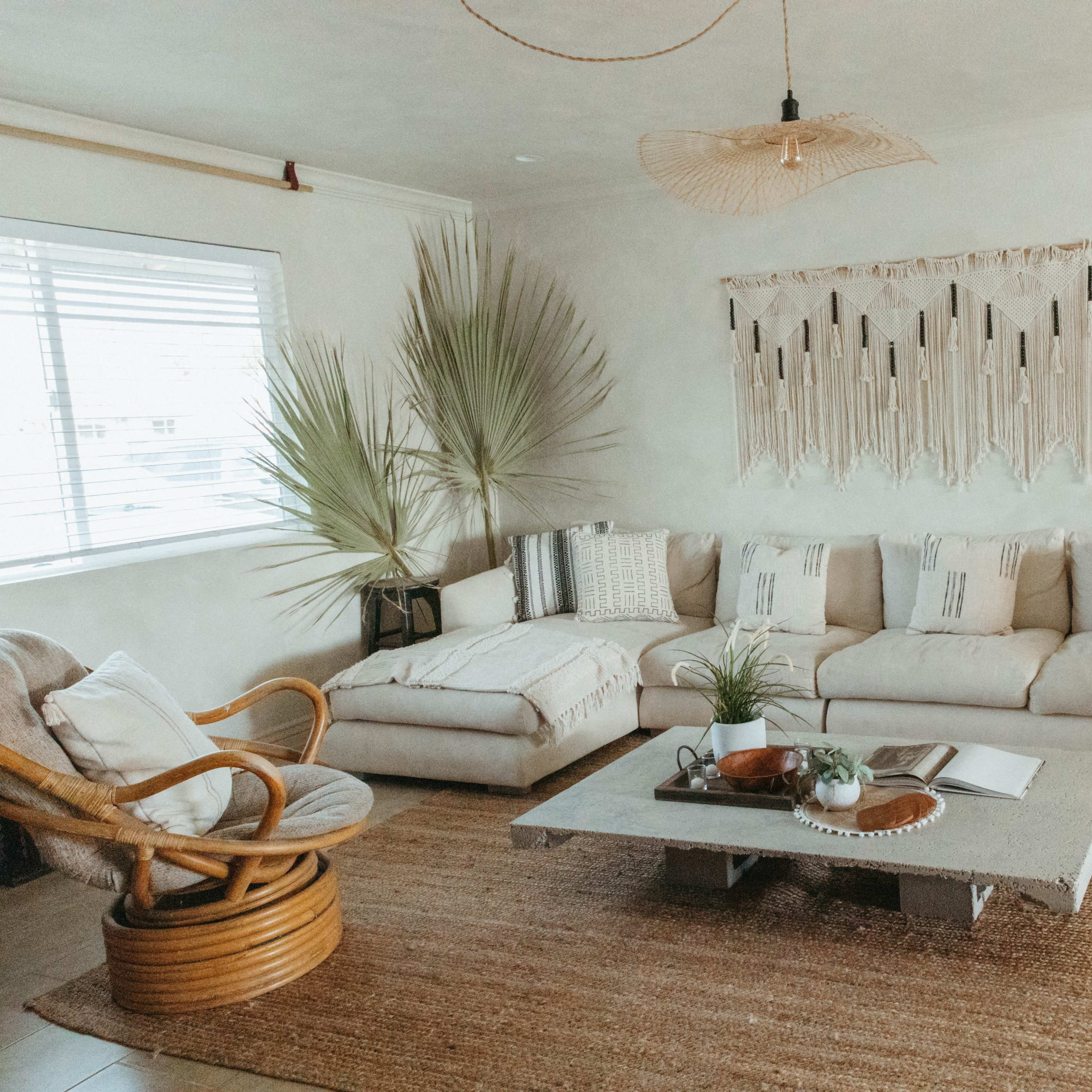 The living room features a large, light-colored sectional sofa, a round rattan chair, and a decorative wall hanging above a coffee table surrounded by plants and a woven rug.