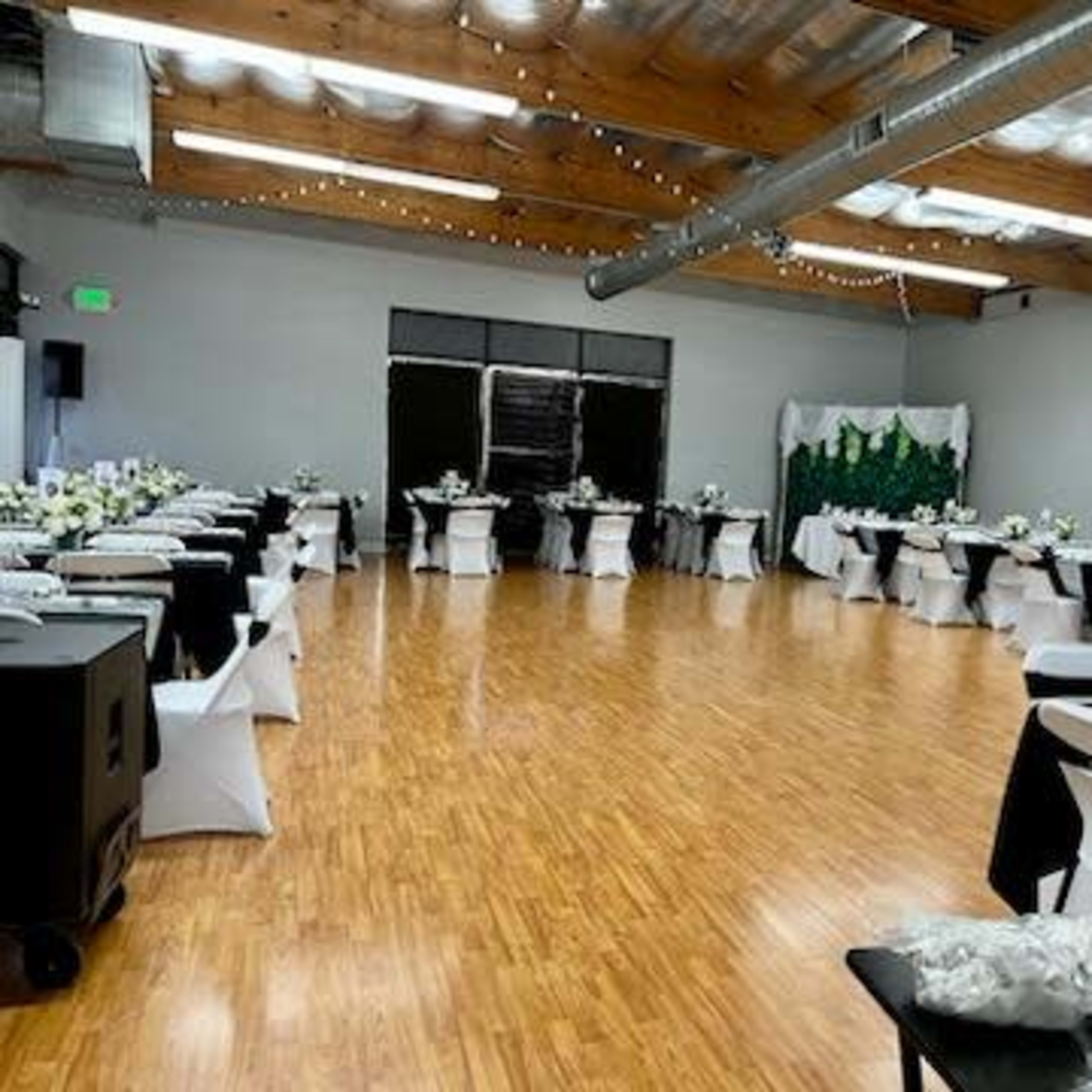 A spacious event hall is set up for a formal gathering, featuring tables covered with black and white linens adorned with floral centerpieces.