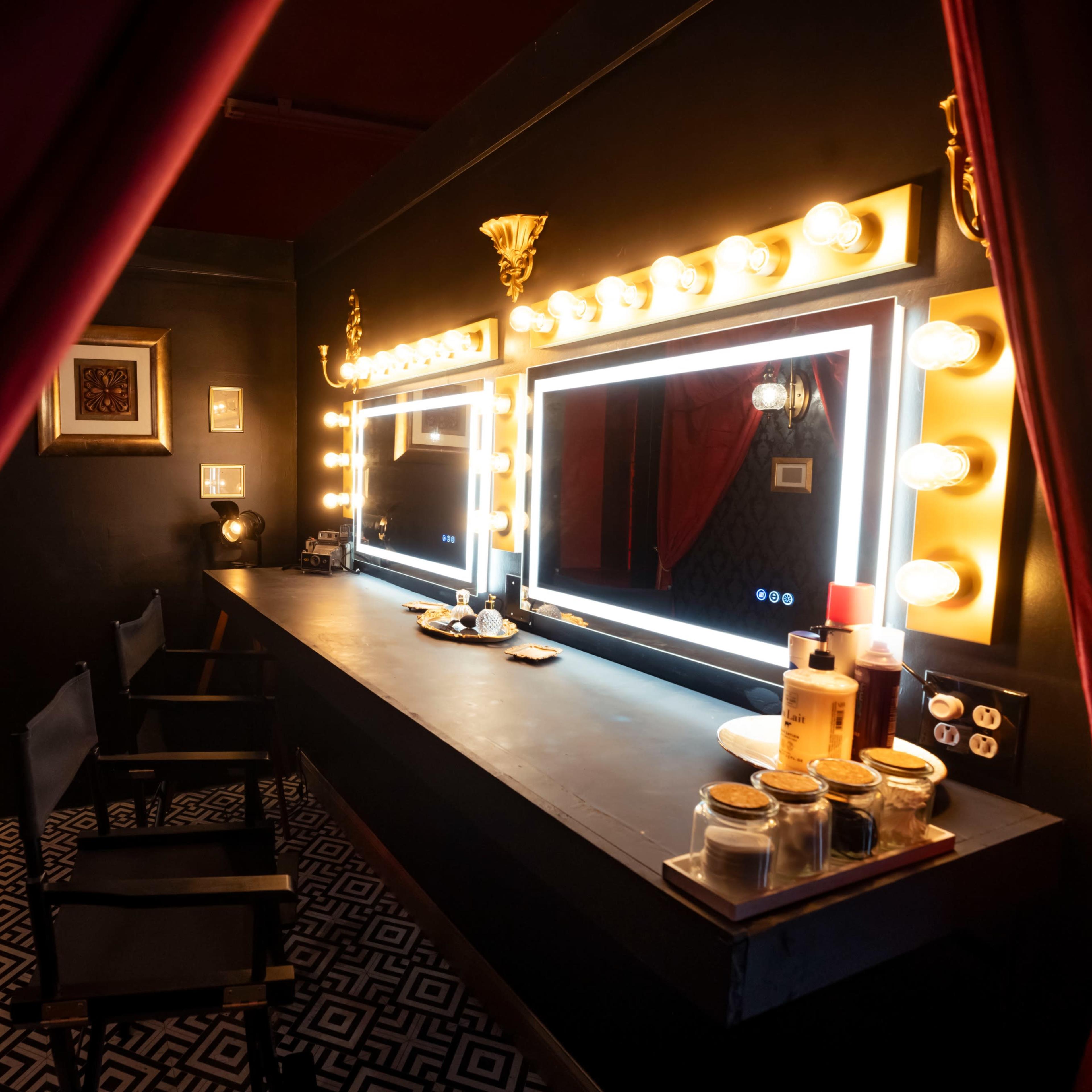 A dimly lit dressing room featuring two large mirrors surrounded by light bulbs, a black counter, and chairs, all set against a patterned floor.