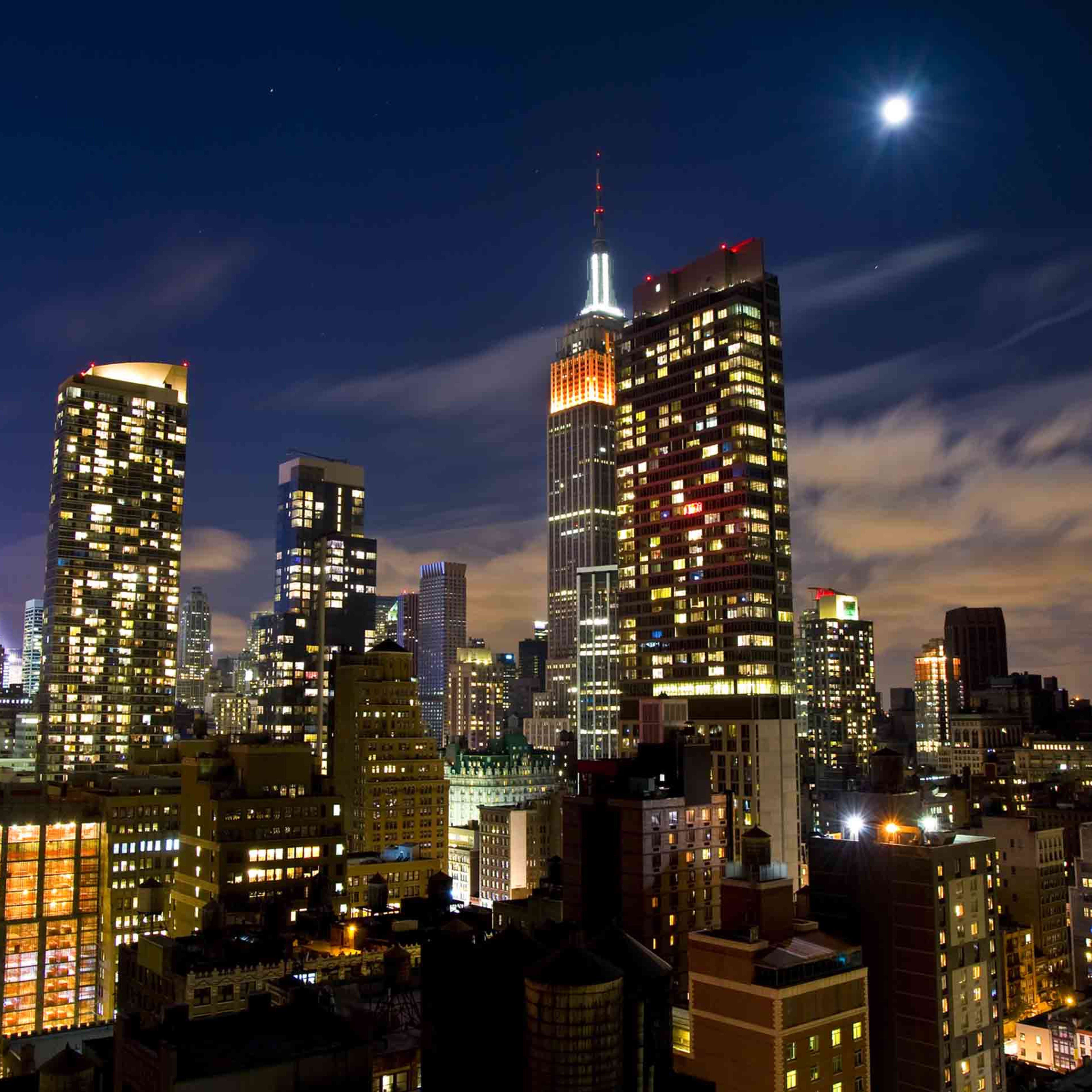 A night cityscape of Manhattan, featuring illuminated skyscrapers and the Empire State Building under a crescent moon.